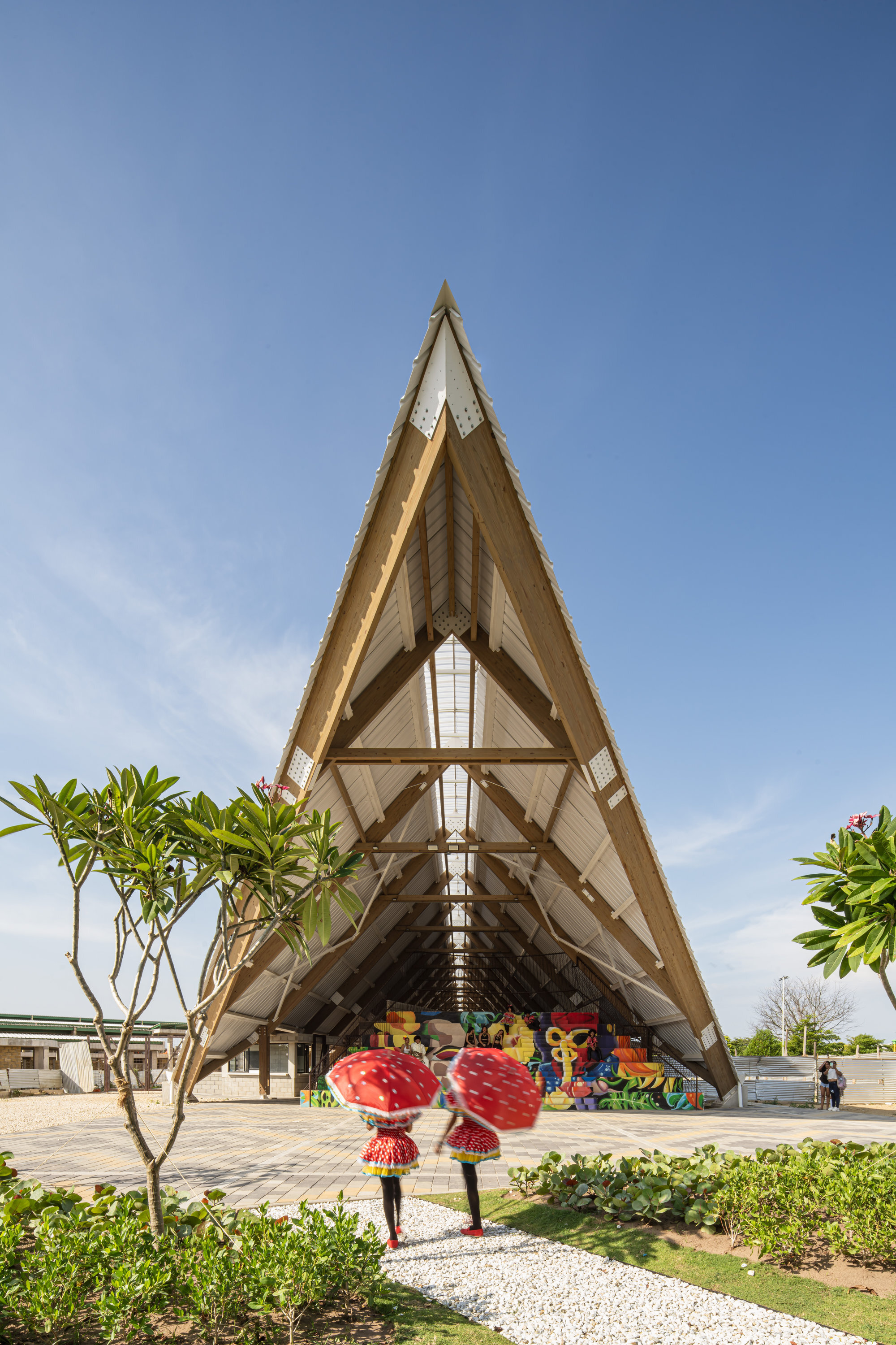 Gable-end view of the triangular timber frame with a mural at the back, as two figures with red umbrellas walk the gravel path
