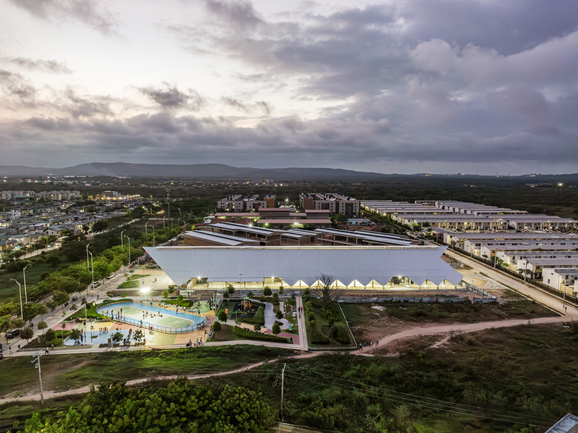 Aerial dusk view of the white pitched roof beside a lit sports court, surrounded by housing blocks under heavy clouds