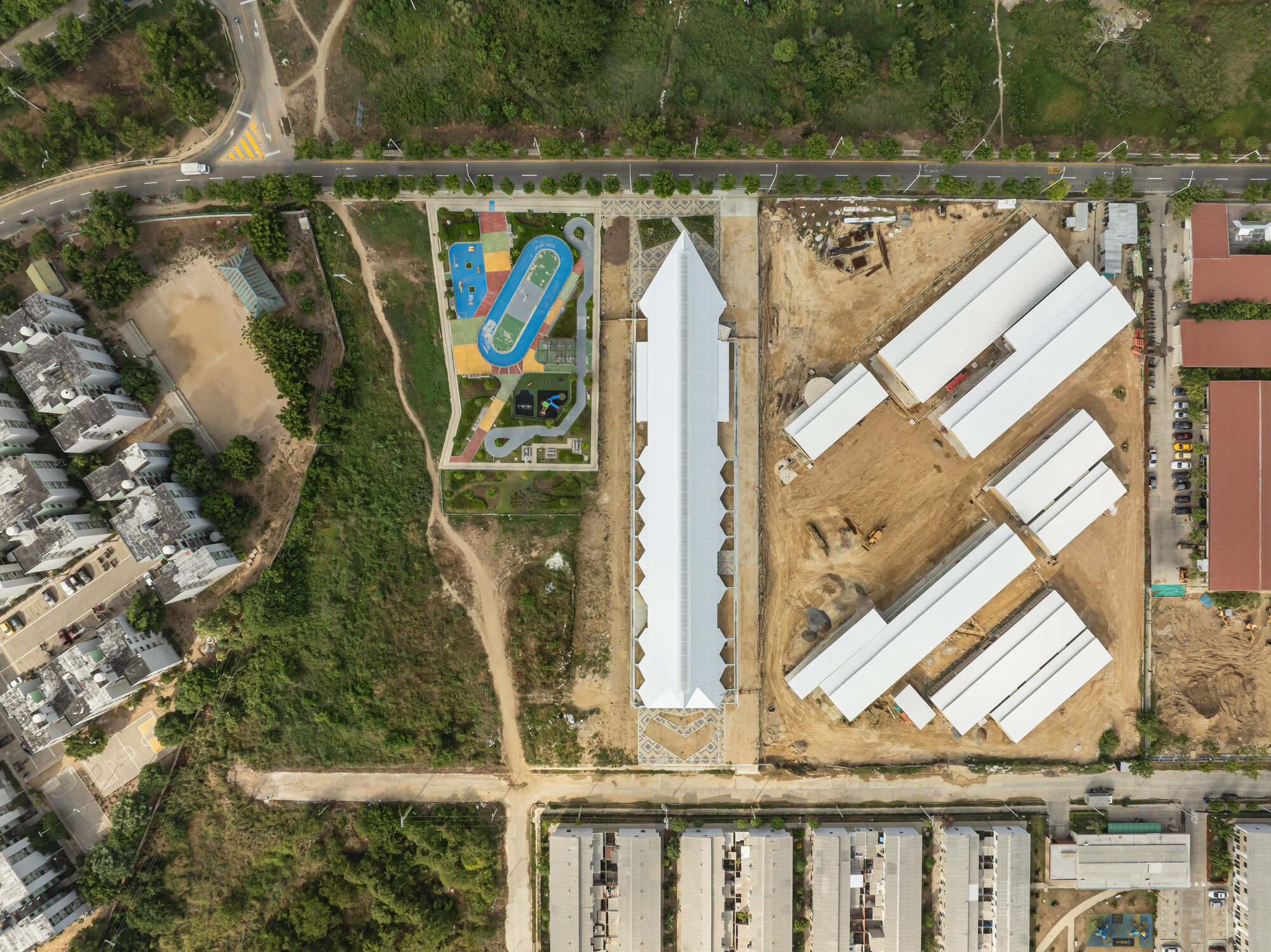 Top-down view of the long white gabled roof between a colorful playground and a dirt construction site with rows of roofs