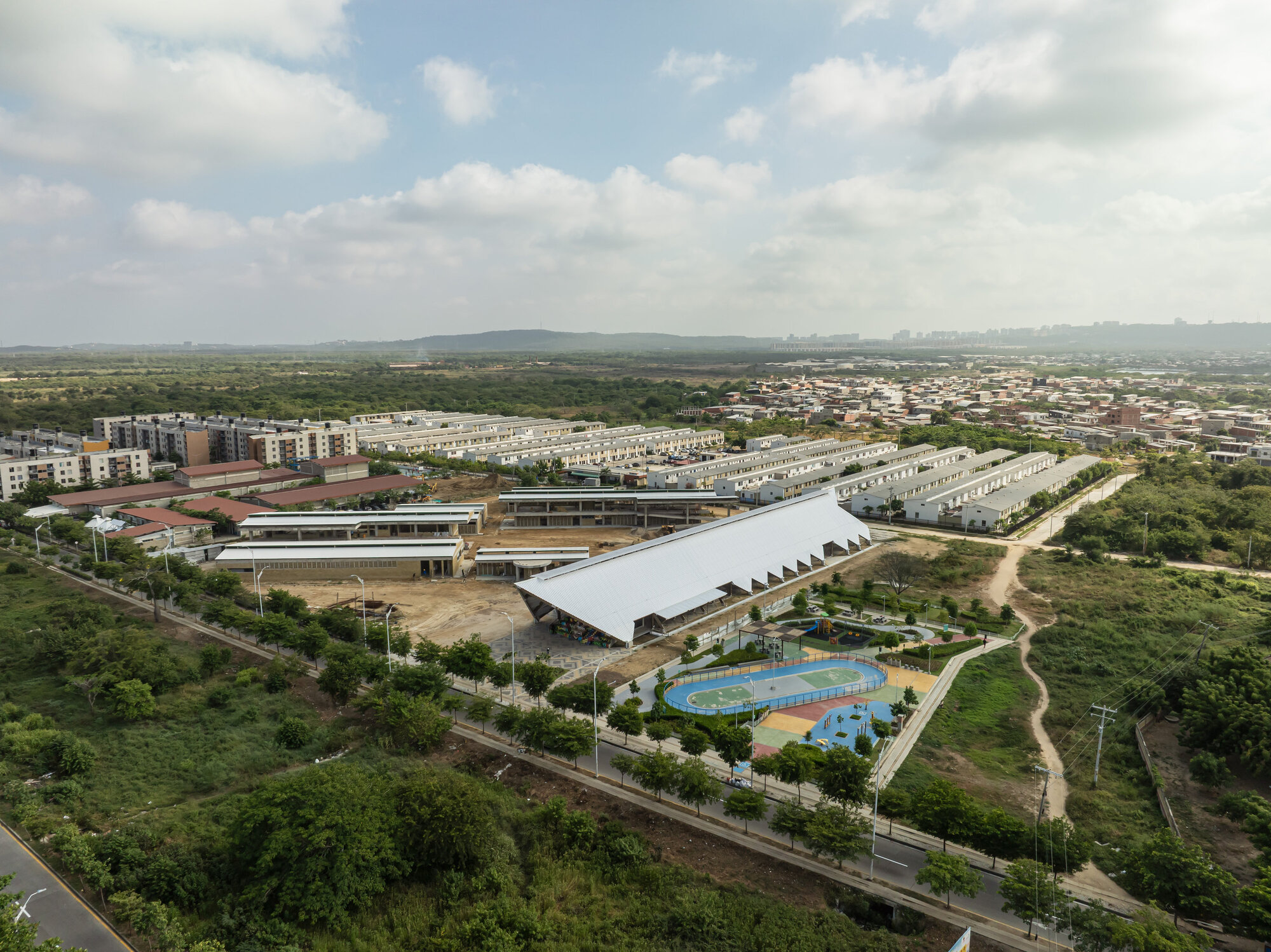 Aerial view of the large white roof beside a blue running track, with housing blocks stretching toward distant hills in daylight