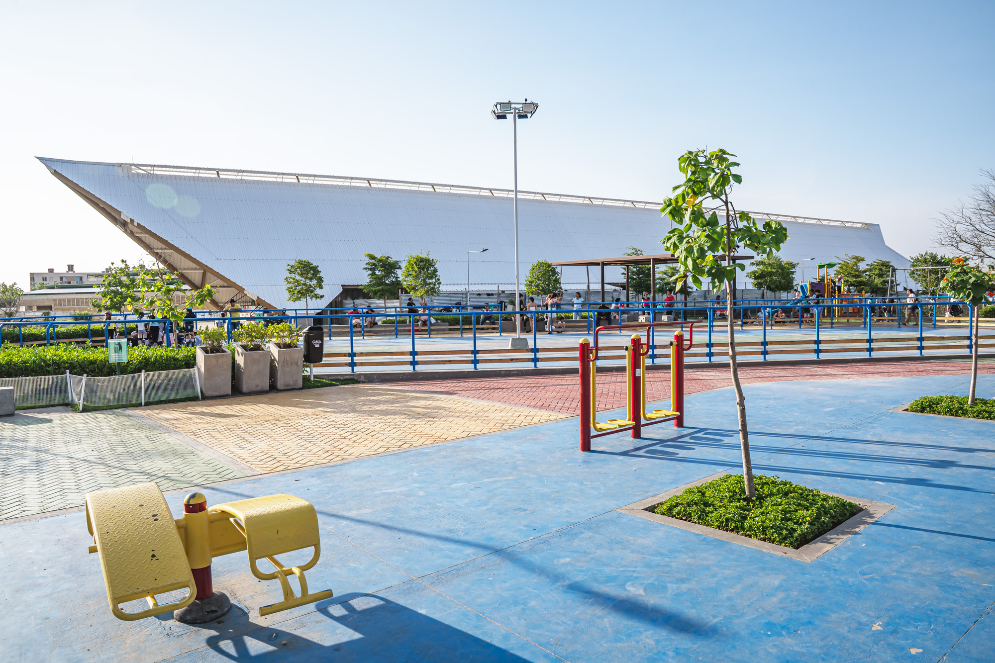 Sloped white roof seen across a blue play court with yellow and red exercise equipment and a young tree in afternoon sun