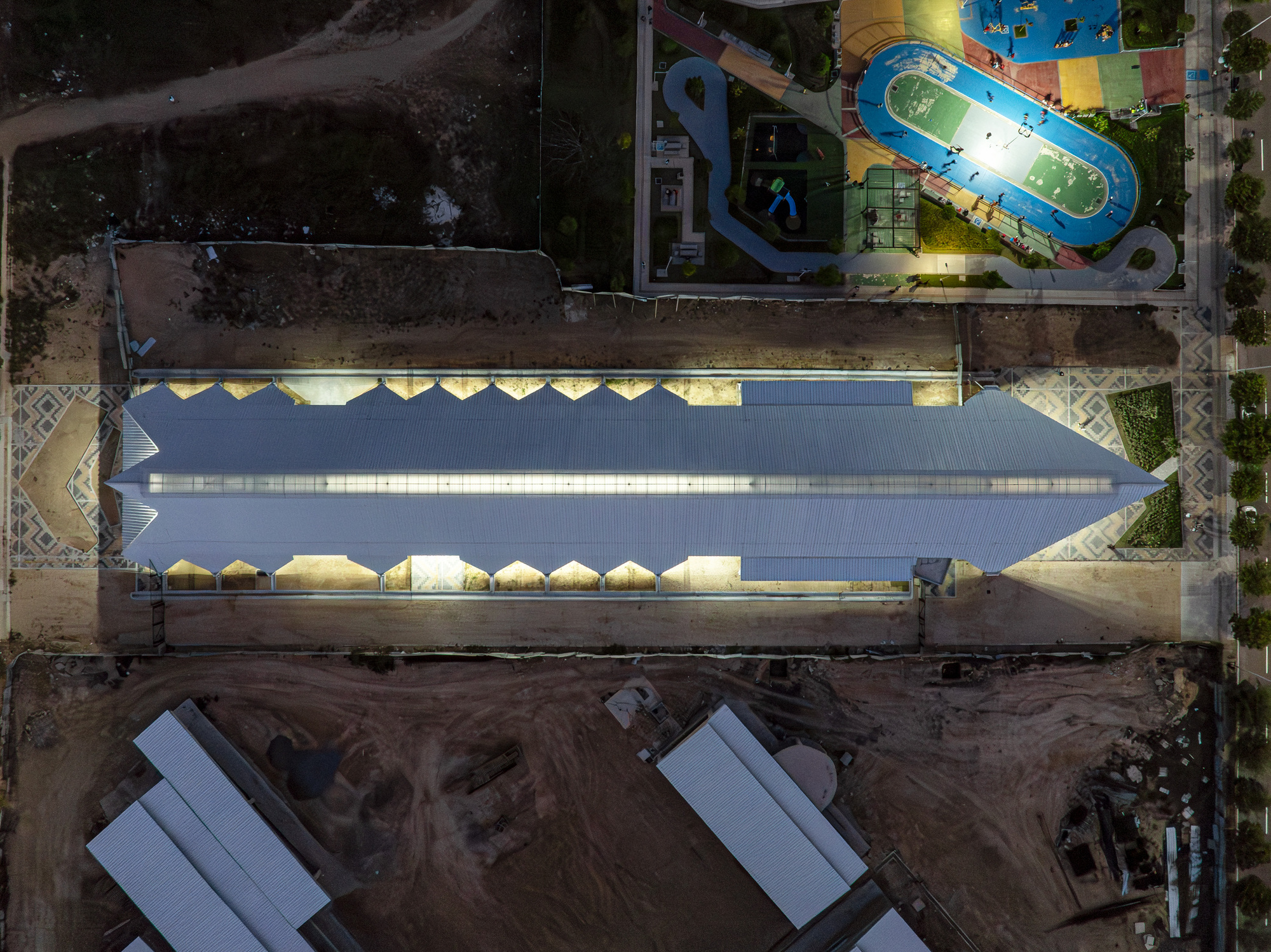 Aerial view of the long corrugated roof with lit zigzag edges, flanked by a colorful sports court at night