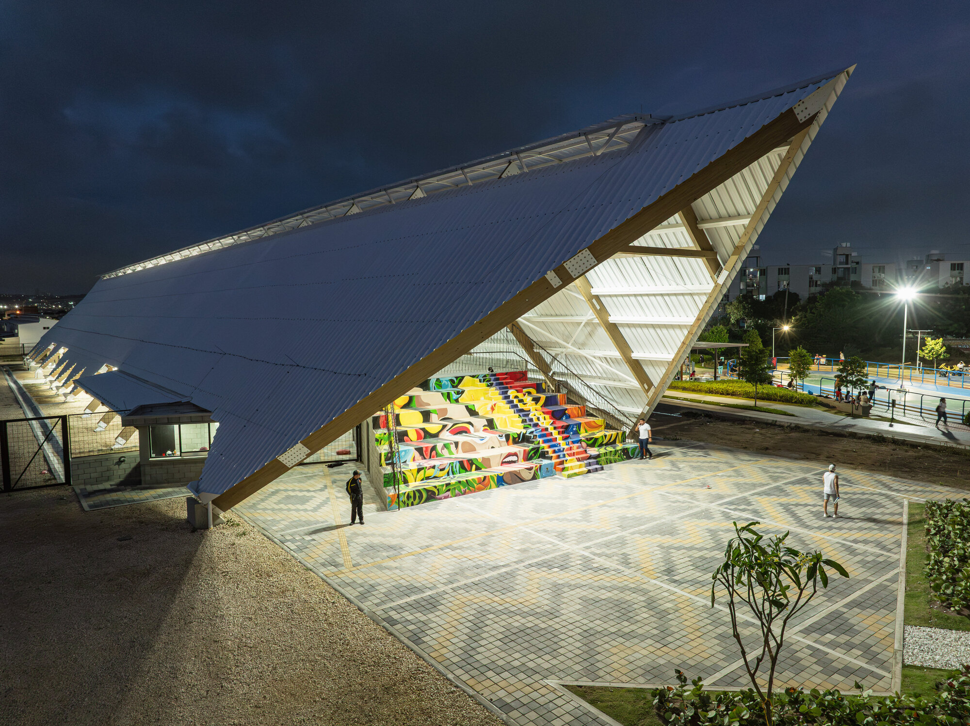 Tilted roof plane over a painted stepped seating wall, with a patterned paved plaza and people under a dusk sky
