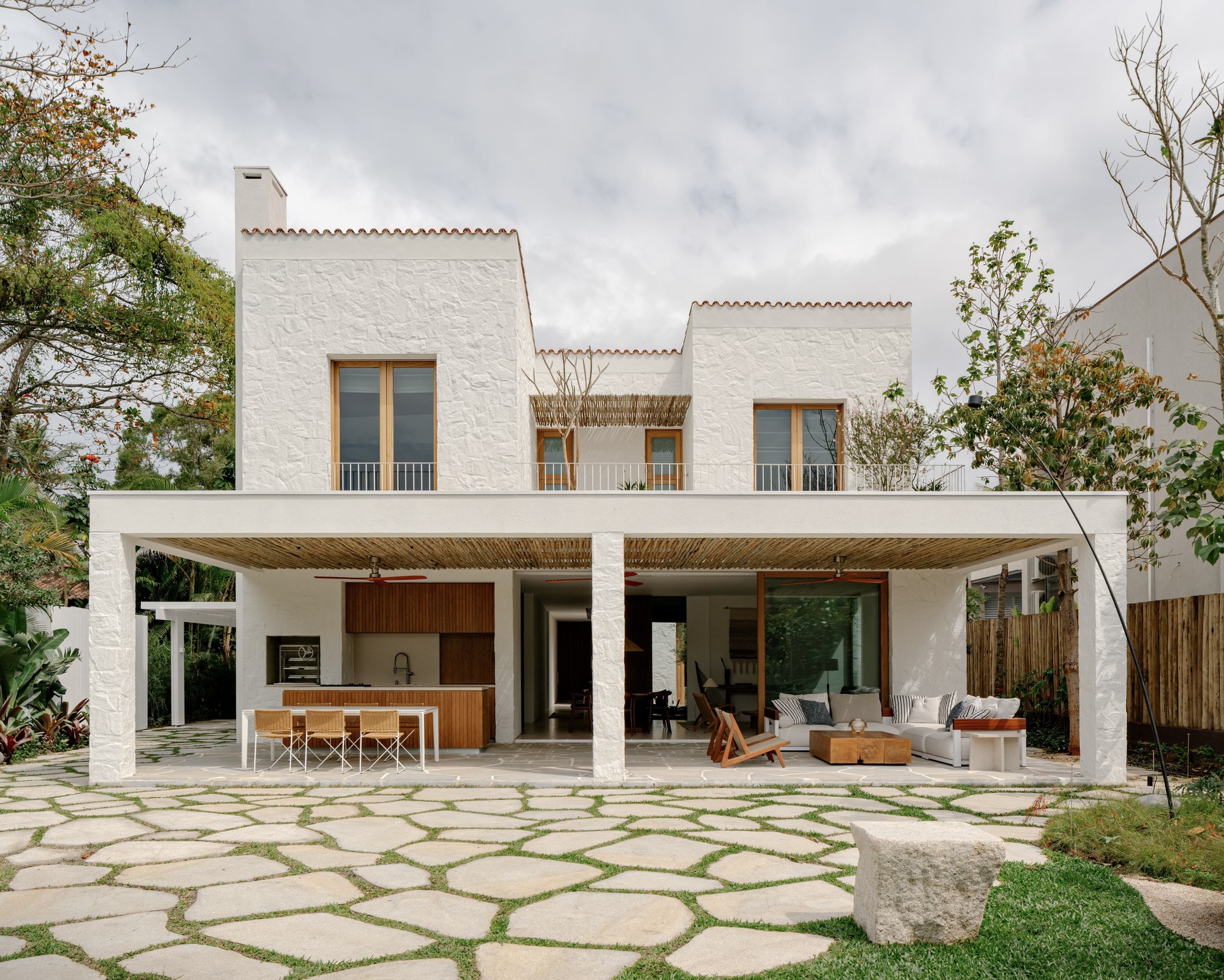 Rear facade of textured white plaster with a deep covered porch, bamboo ceiling and irregular stone paving under an overcast sky