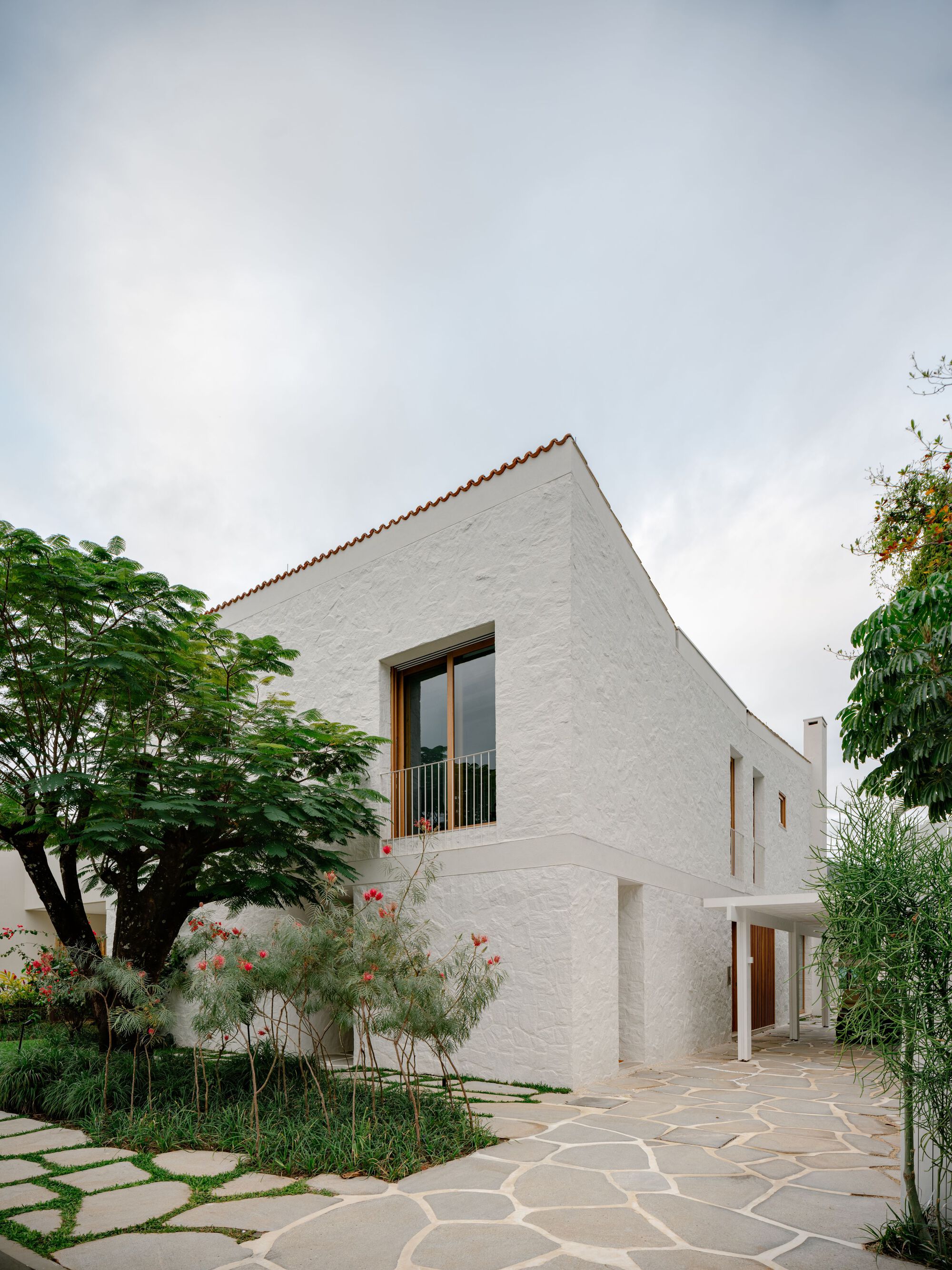 Corner of the textured white plaster volume with a timber-framed window, clay tile edge and flowering shrubs beside a stone path