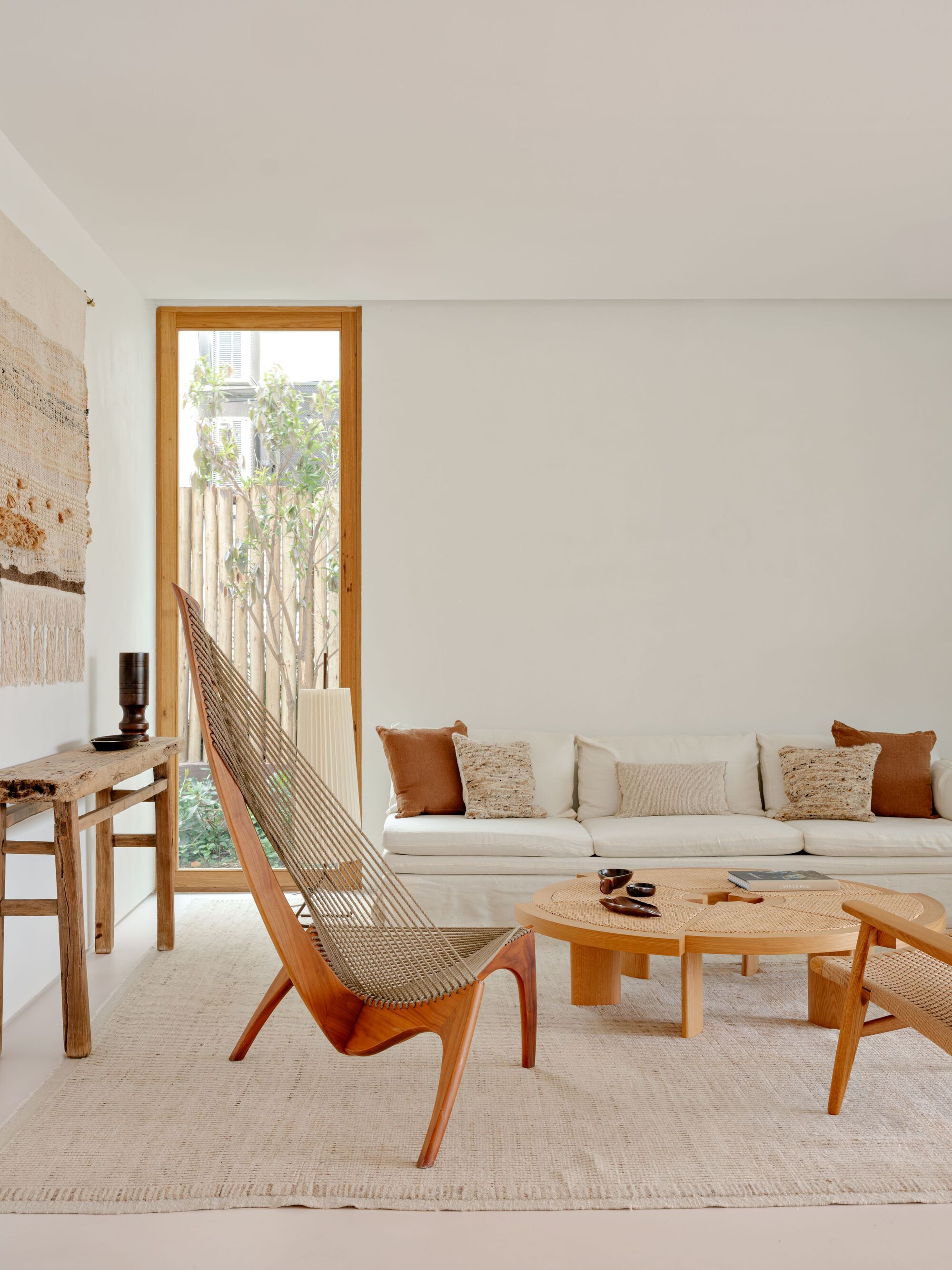 Tall timber-framed window beside a white linen sofa, a sculptural rope lounge chair and a round wood coffee table