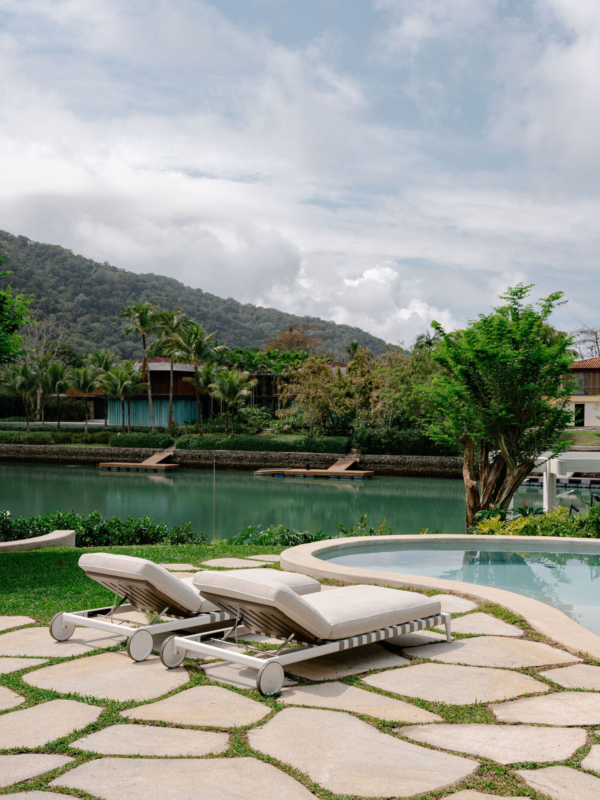 Two cushioned loungers on stone paving beside a curved pool edge, facing a green lagoon and forested hill under clouds