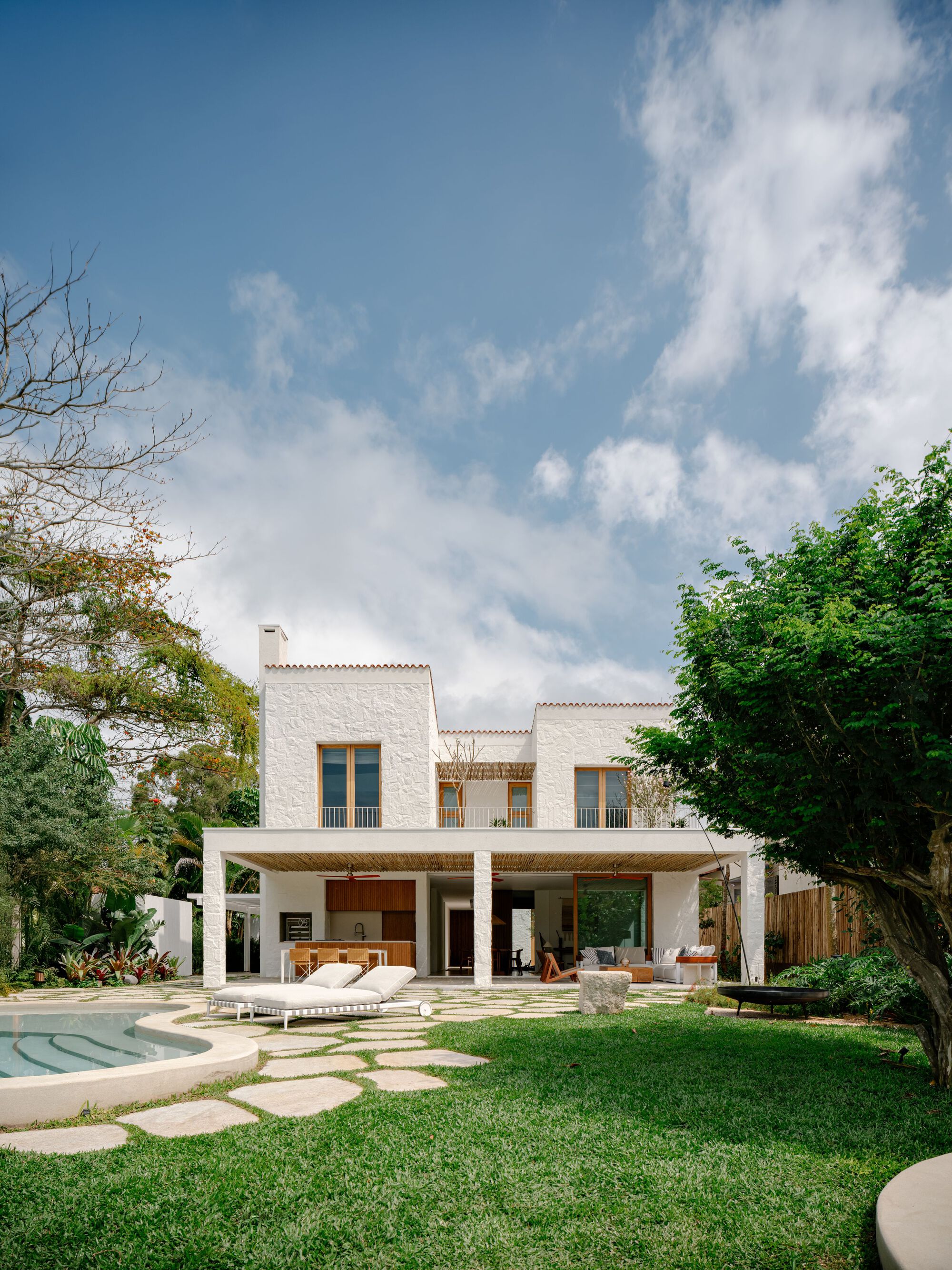 Two-storey white house seen across a lawn, with a pool at left, loungers on stone paving and a leafy tree at right