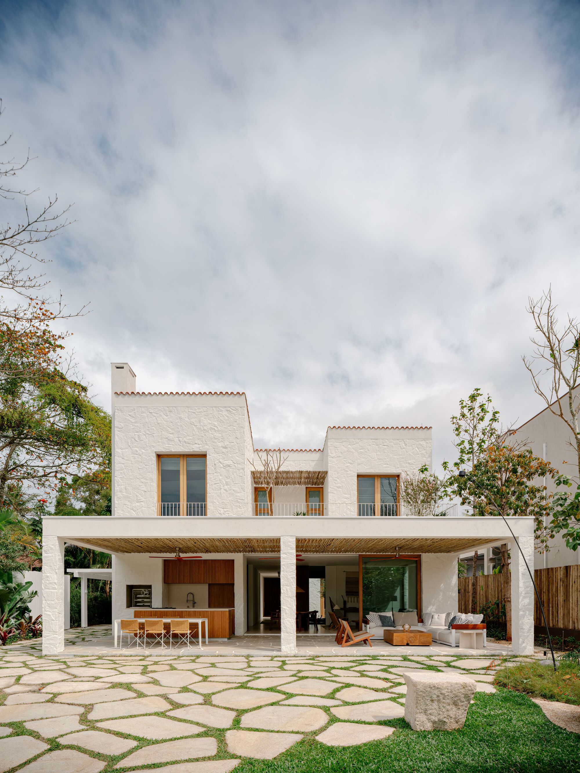 Rear facade of the two-storey white house with a covered porch, timber-framed windows and irregular stone paving under a cloudy sky