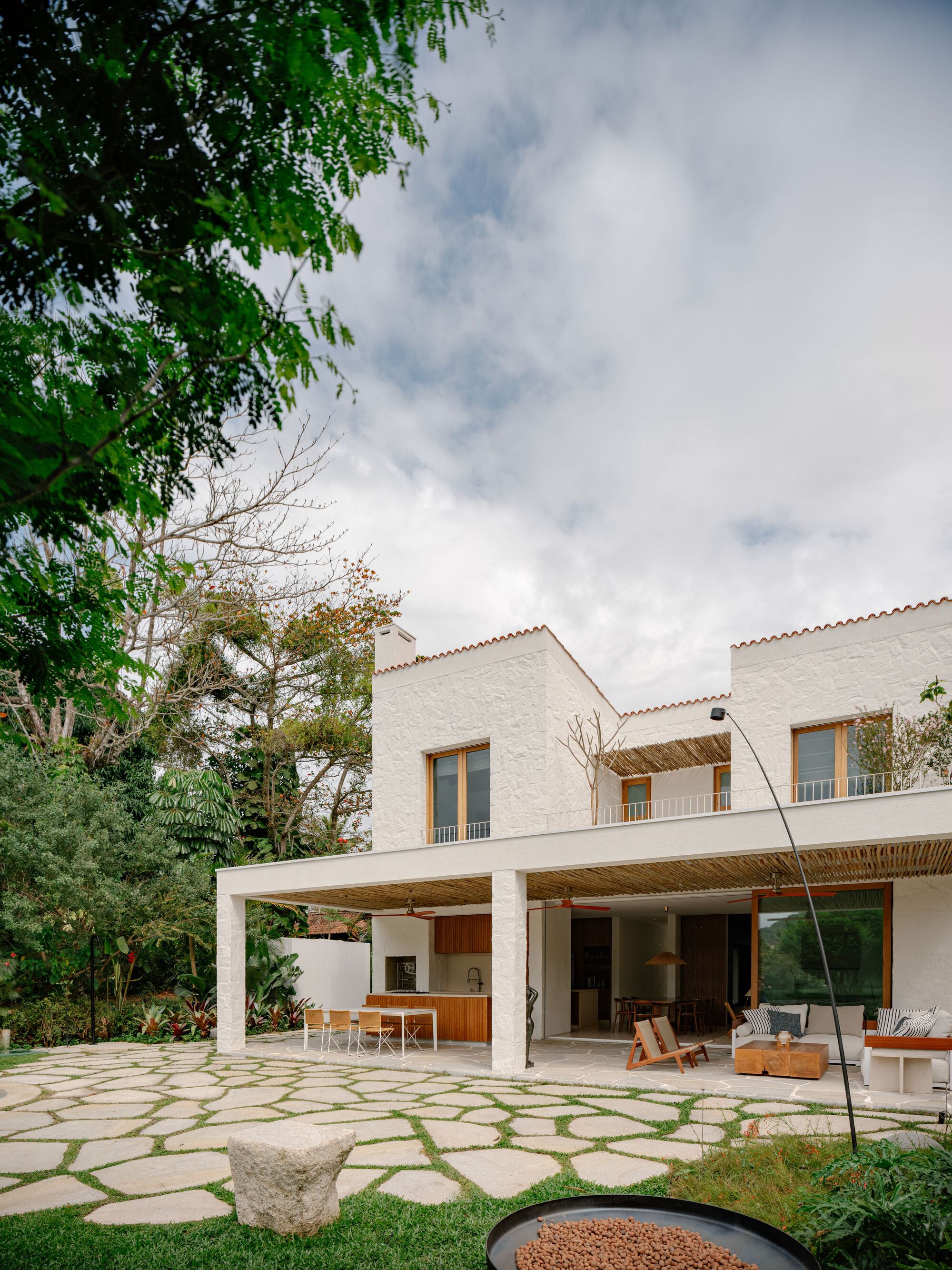 Angled view of the textured white house and open ground-floor porch, framed by tree foliage and a flagstone terrace