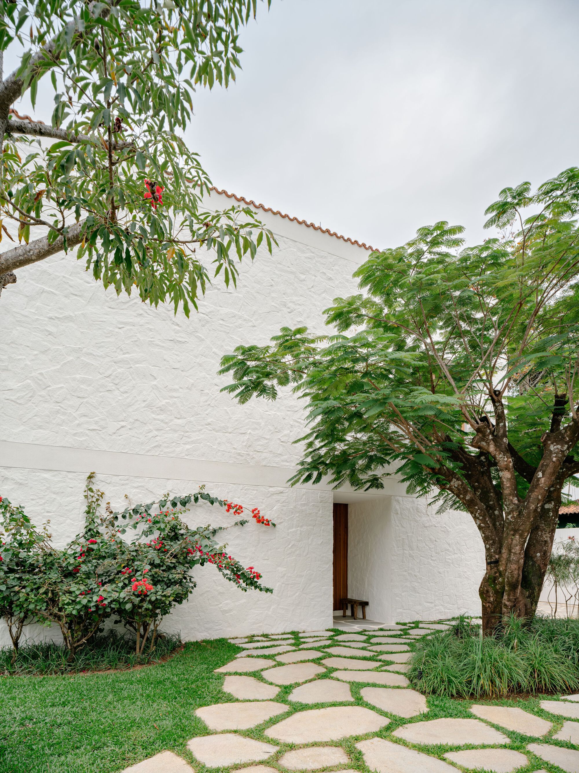 Recessed timber entrance door in a textured white wall, with a flagstone path, red-flowered shrub and tree canopy on an overcast day