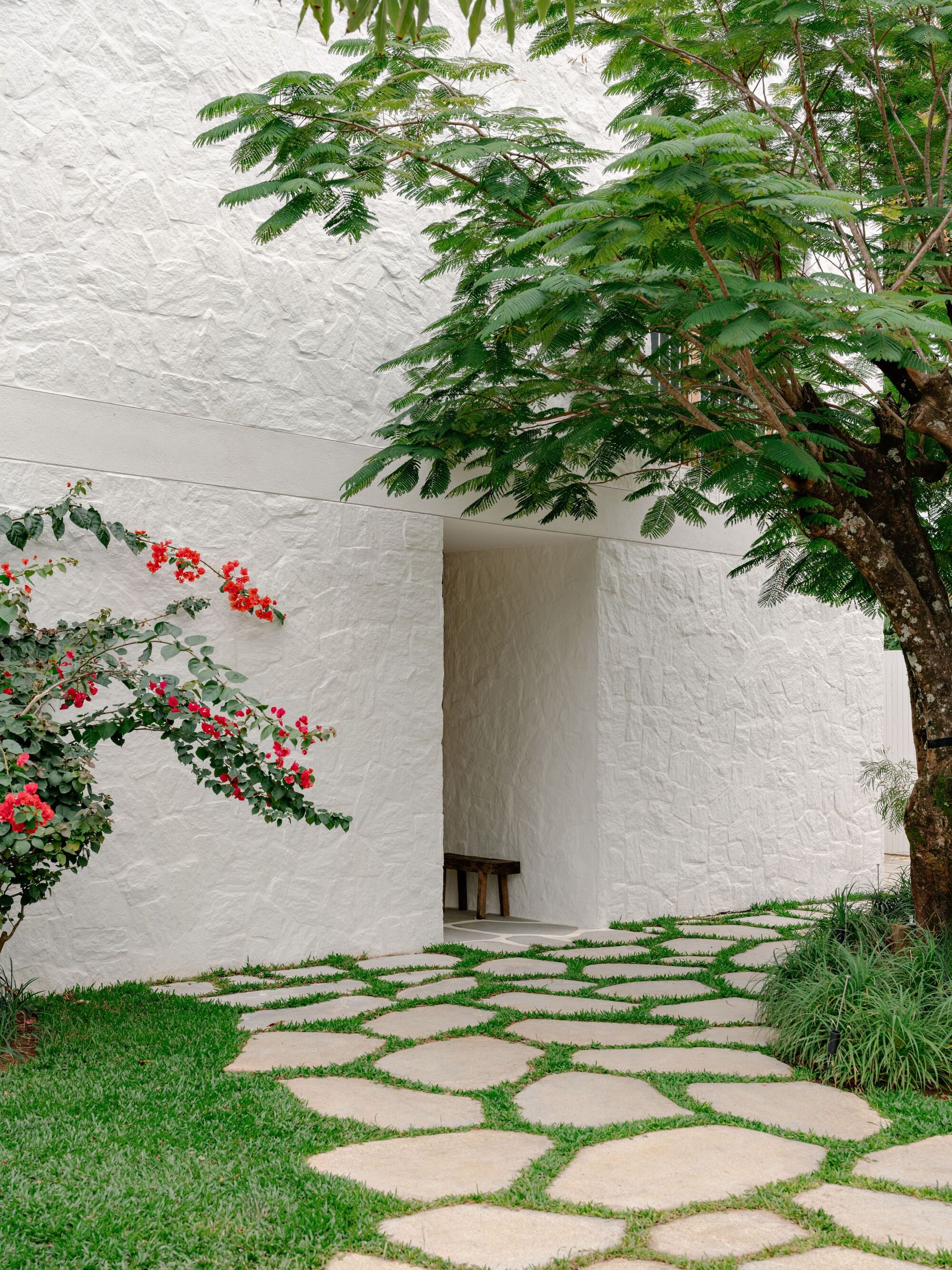 Textured white plaster wall with a recessed entry and wooden bench, beside a flowering bougainvillea and irregular stepping stones set in grass