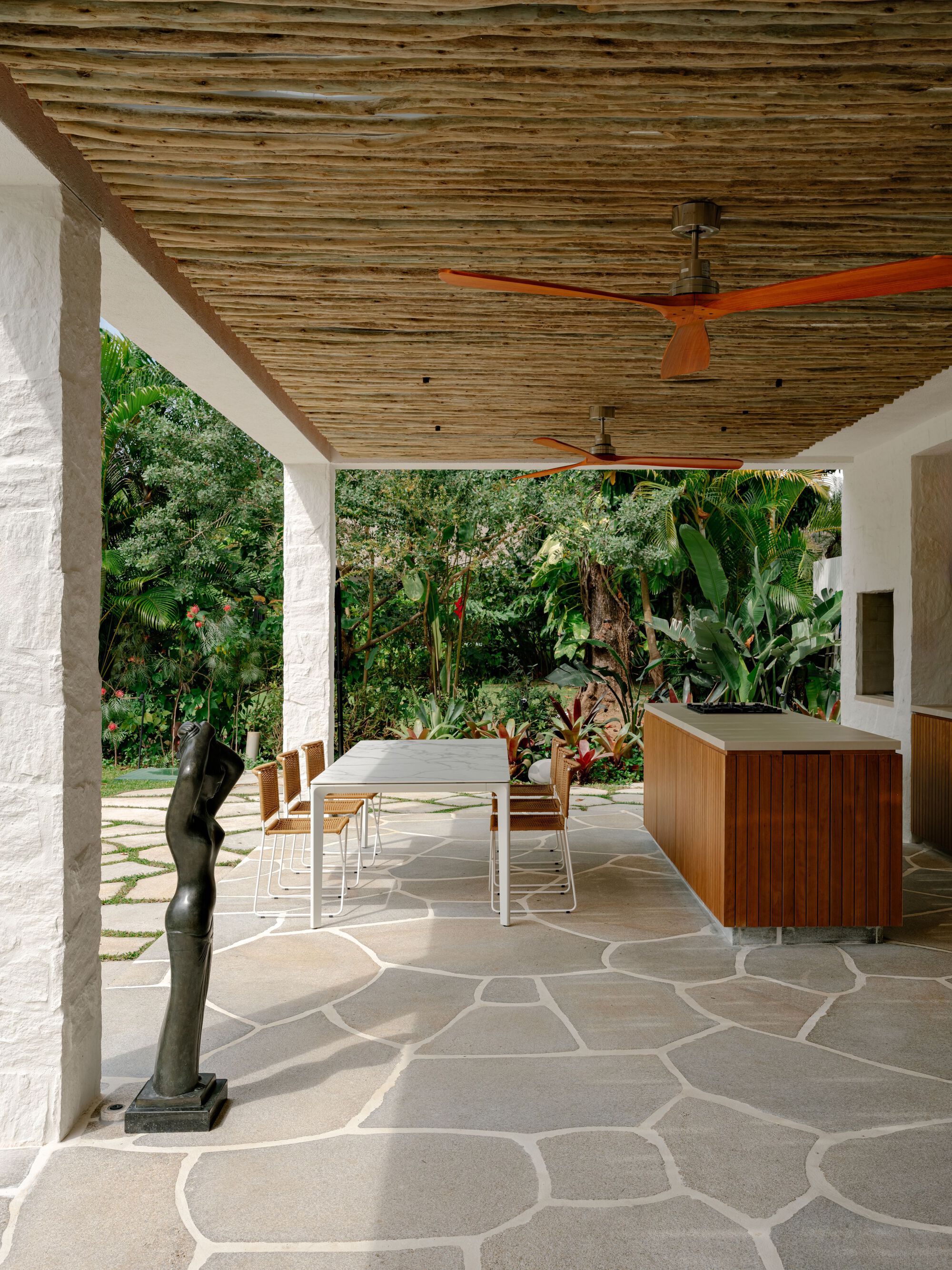 Covered terrace with a reed ceiling, orange fan blades, dining table and timber kitchen island facing a lush garden