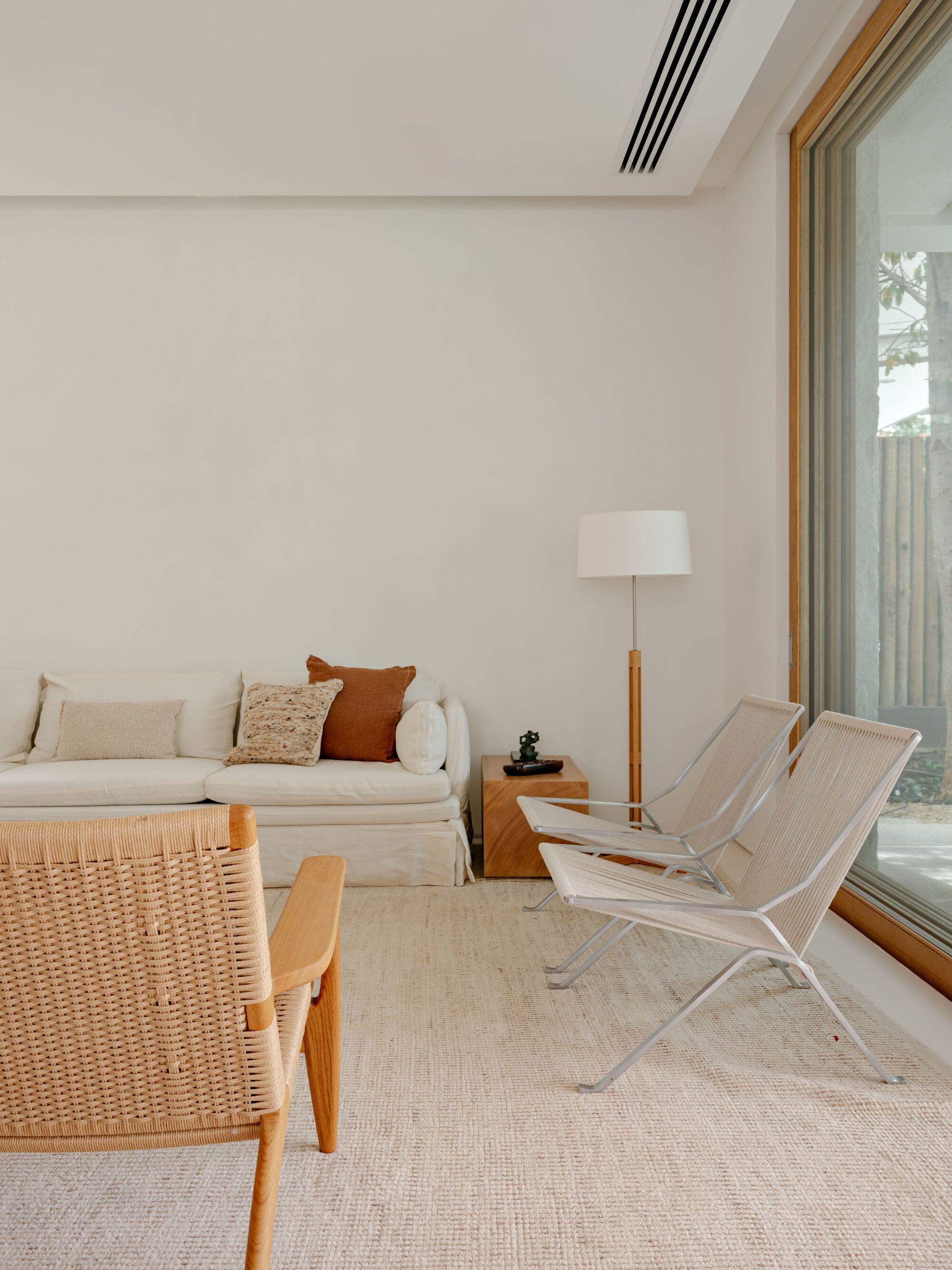 Living room corner with a linen sofa, rush-seat armchair and steel-framed lounge chairs beside a tall timber-framed window