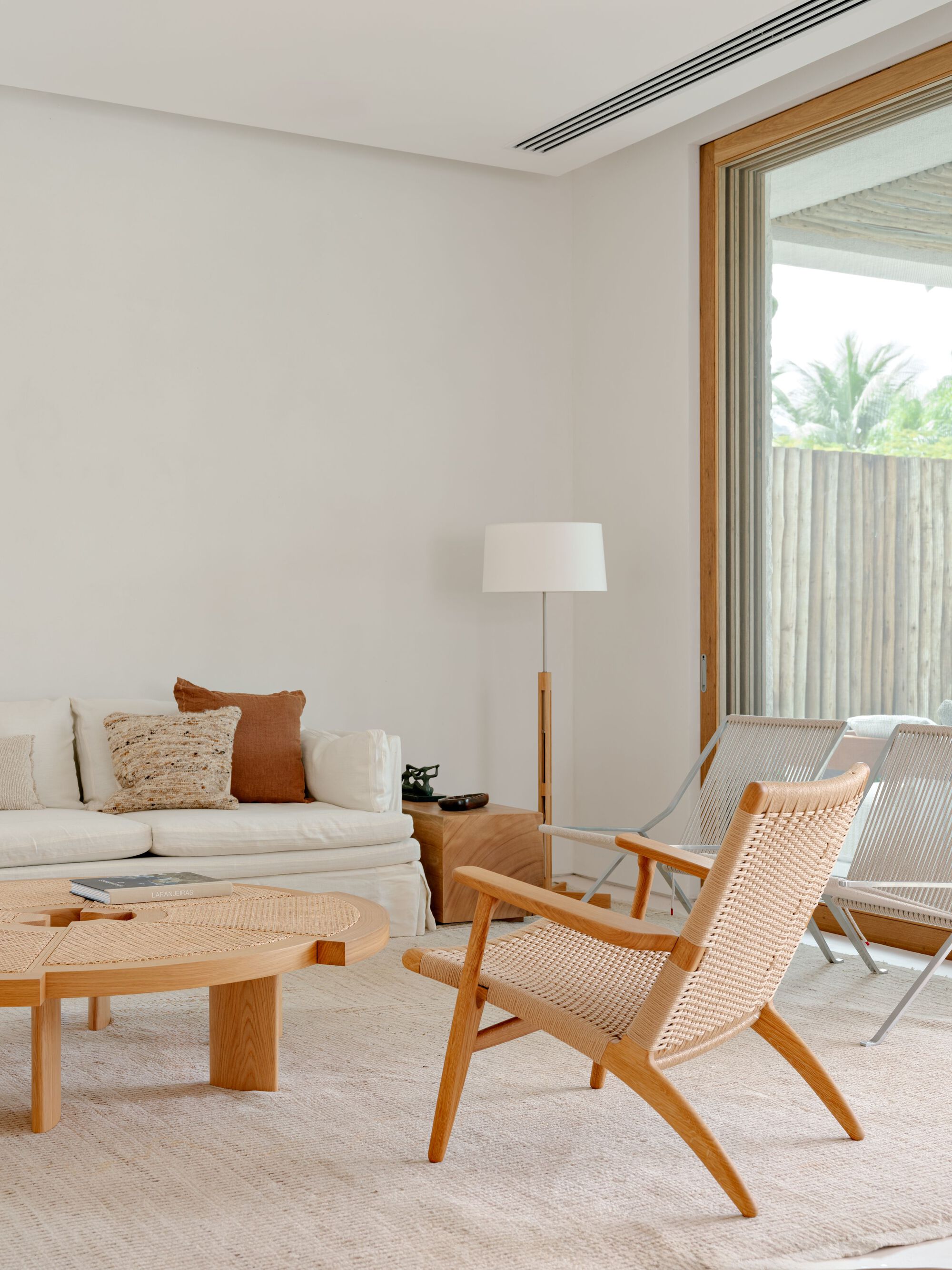 Woven cord lounge chair and round wooden coffee table before a linen sofa, with palms visible through the glazing in daylight