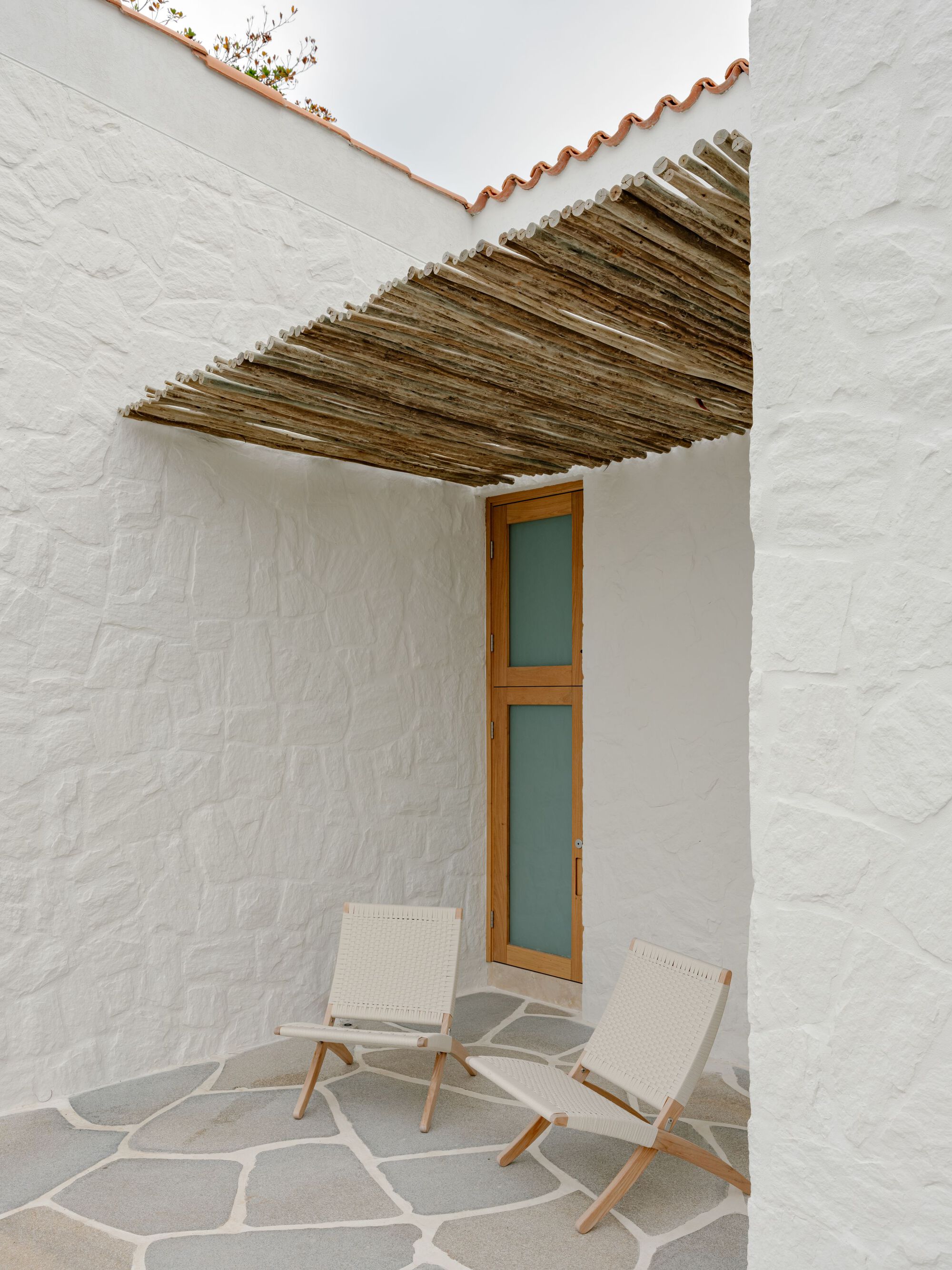 Whitewashed courtyard corner with a reed lattice canopy, tall teal-glazed timber door and two woven chairs on irregular flagstone paving