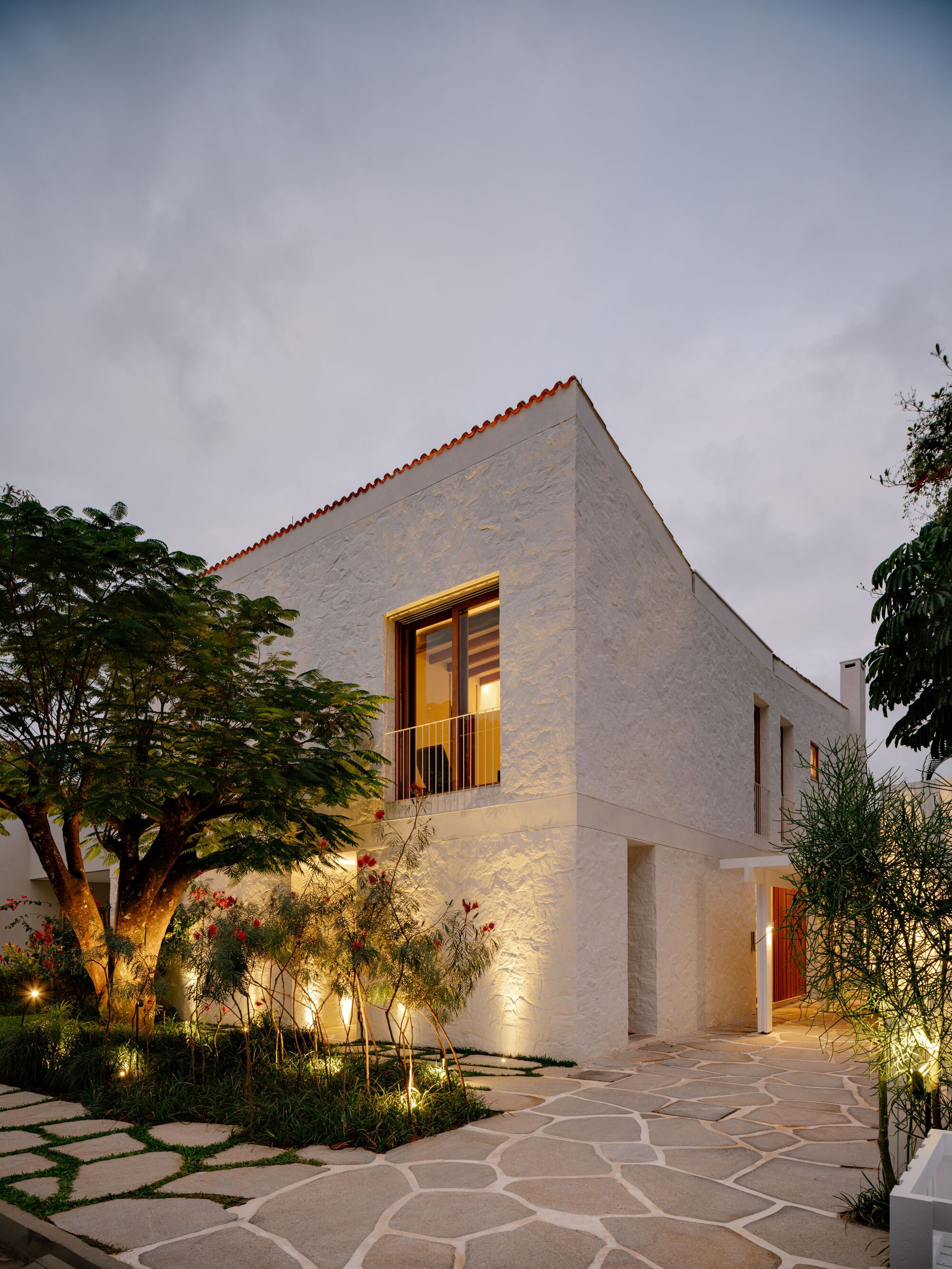 Rough white stone house corner at dusk, lit window with balcony rail, uplit planting and flagstone path beneath a cloudy sky