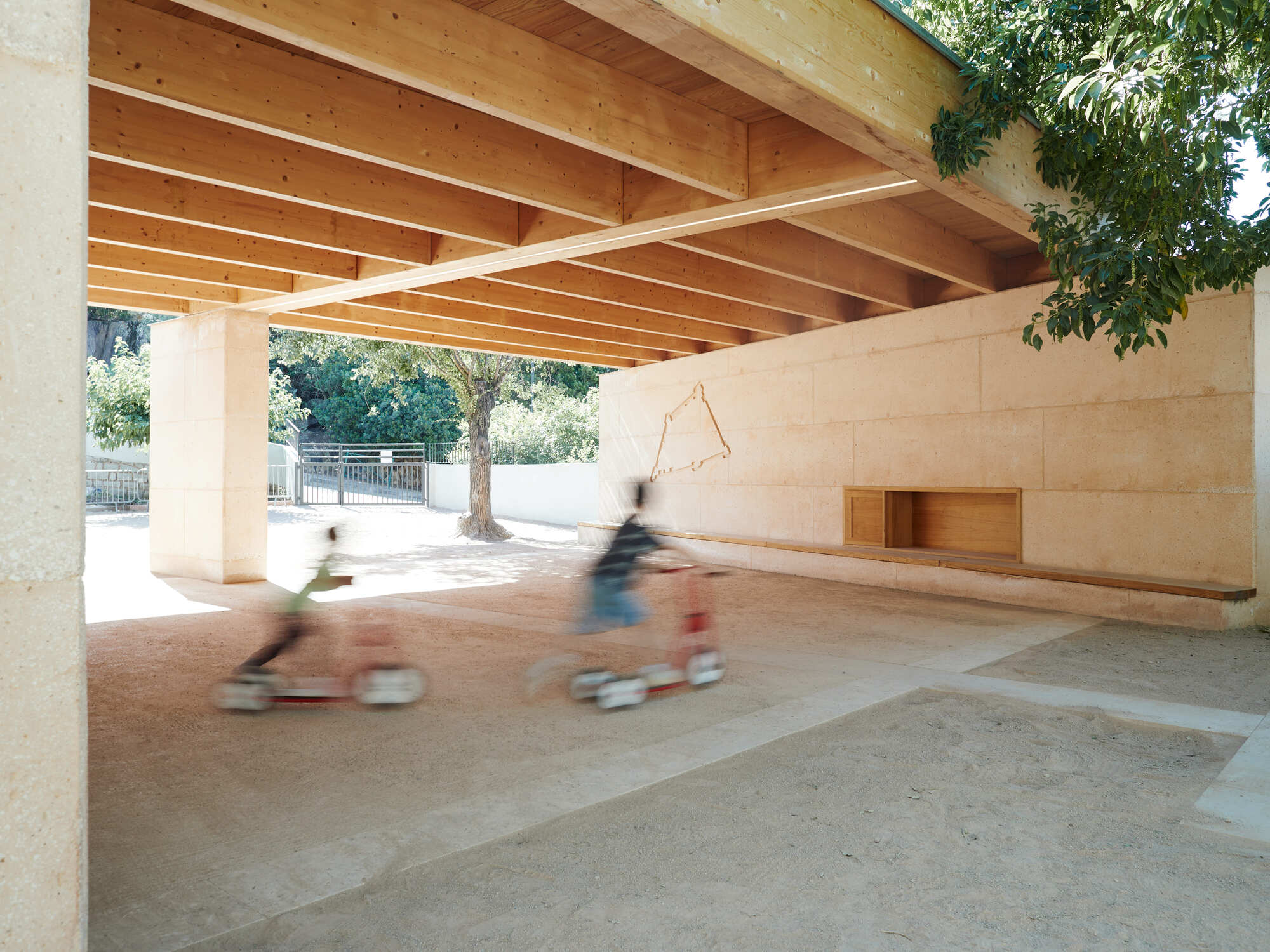 Covered play area with timber beams over a rammed earth wall, as two children on scooters blur past in sunlight
