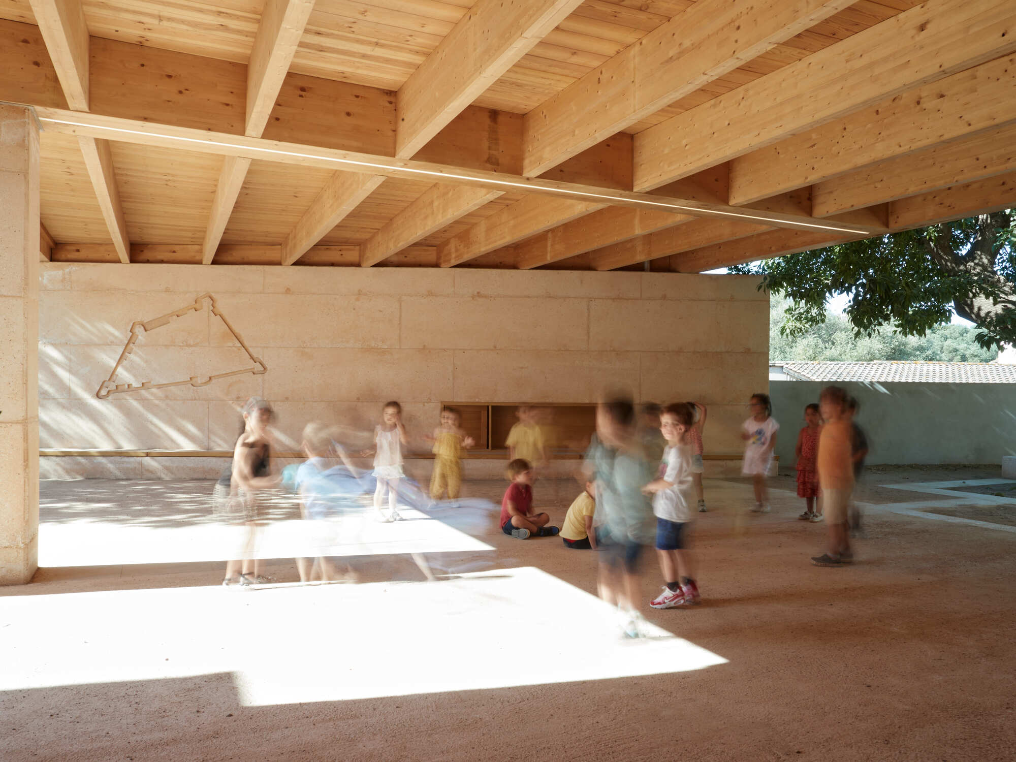 Children playing under a timber beam roof beside a rammed earth wall, with a bright patch of sun on the gravel floor