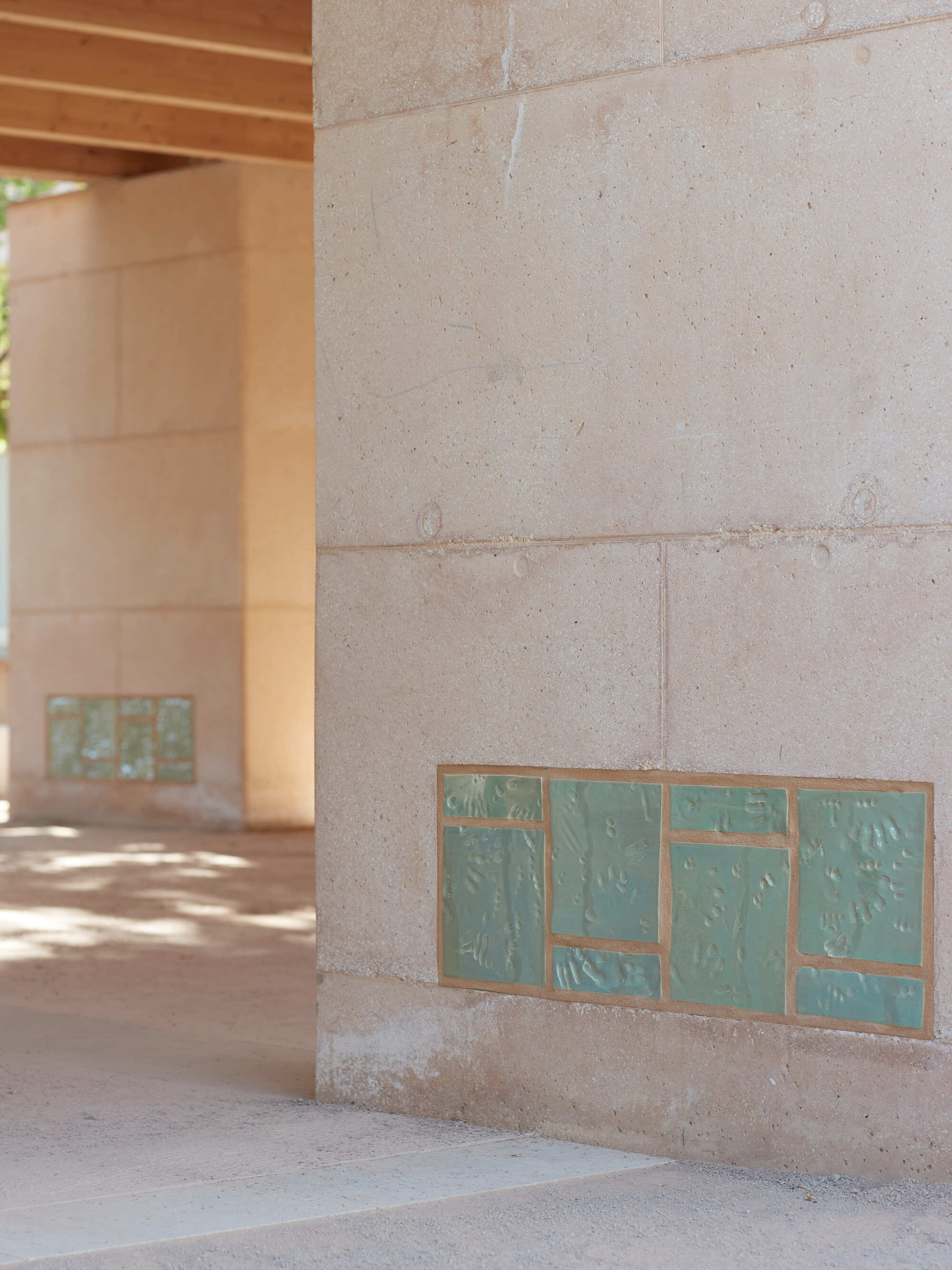 Green glazed ceramic tile panels set into rammed earth columns, with dappled light on the paving beyond