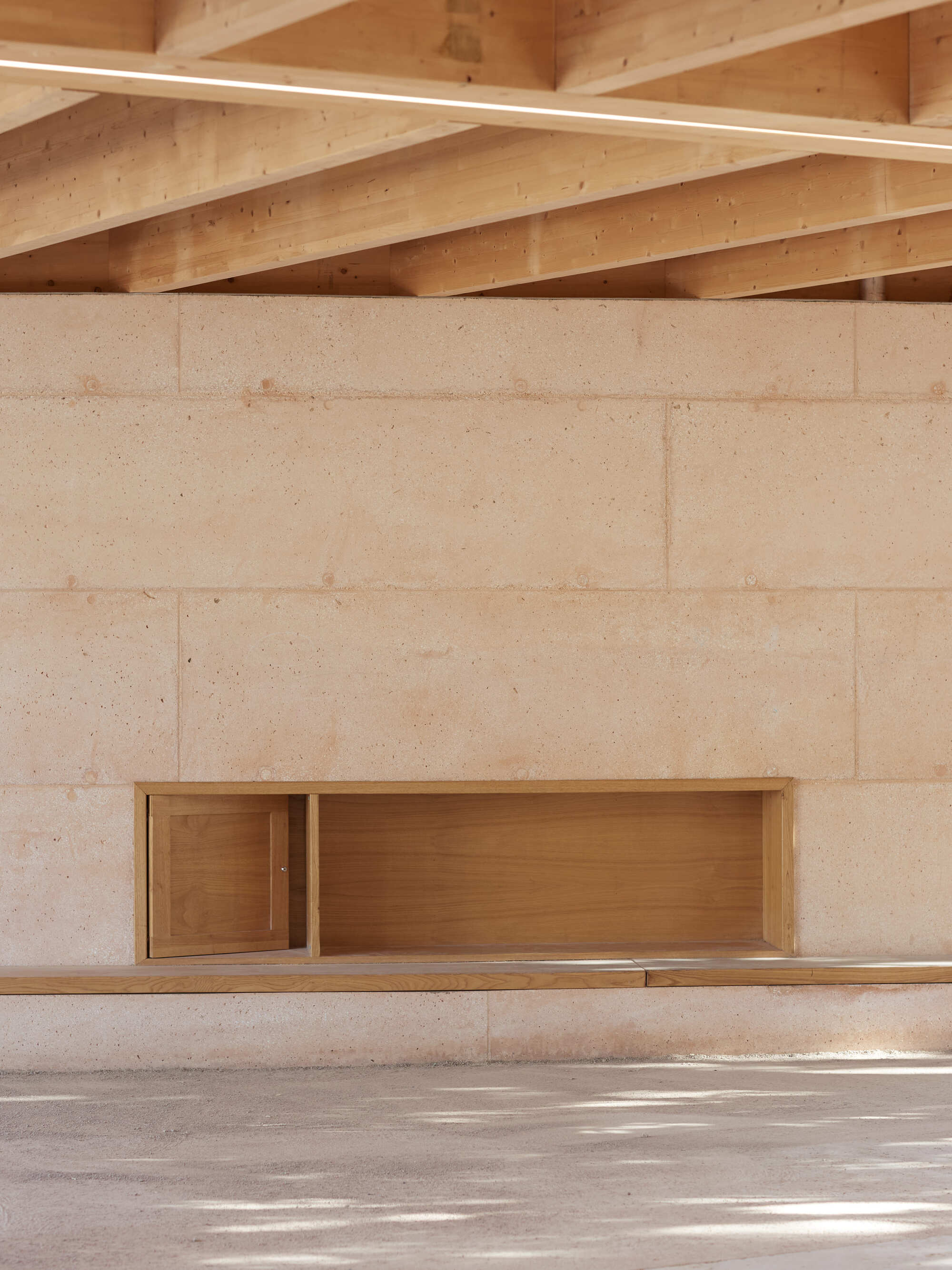 Rammed earth wall with a recessed timber cabinet and built-in bench beneath exposed beams, with leaf shadows on the floor