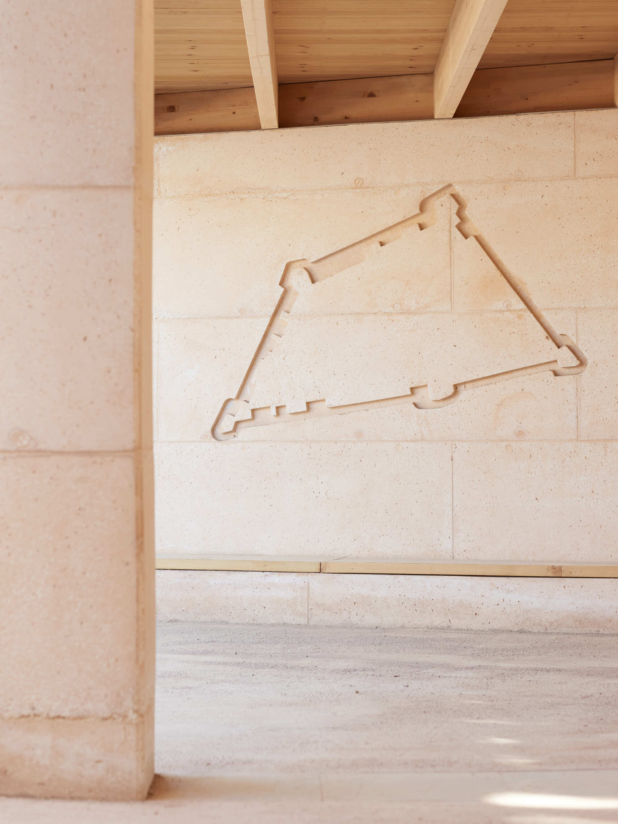 Triangular fortress outline carved into a rammed earth wall above a timber bench, framed by a stone column