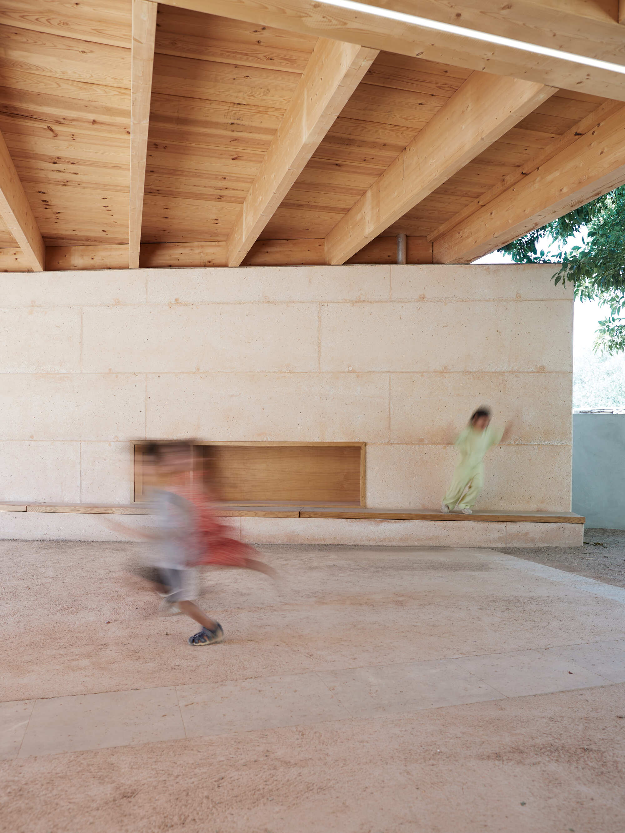 Rammed earth wall with a timber niche and bench under glulam beams, as a blurred child runs across the gravel