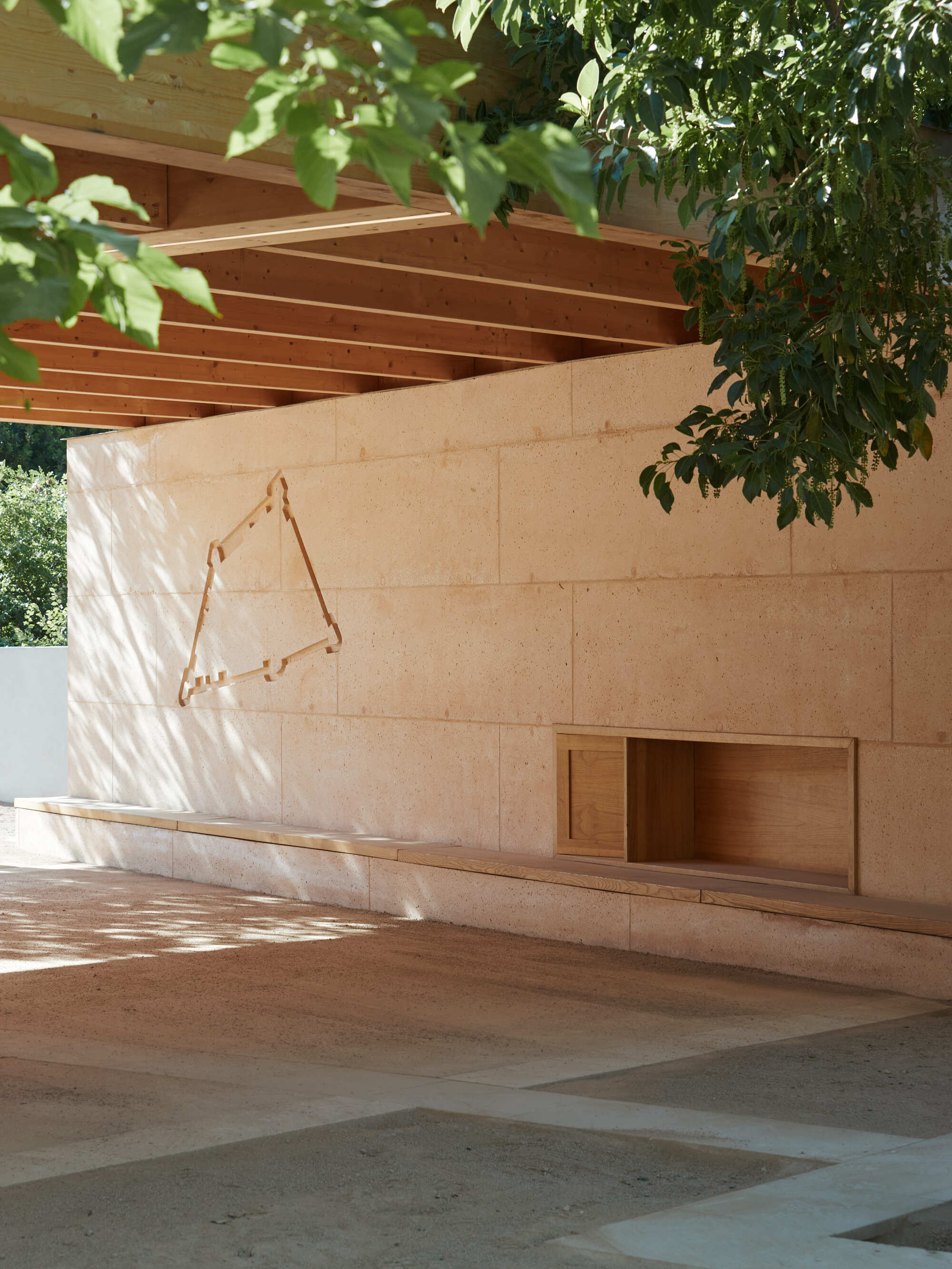 Pale rammed earth wall with a built-in timber niche and bench beneath exposed rafters, dappled by leaf shadows