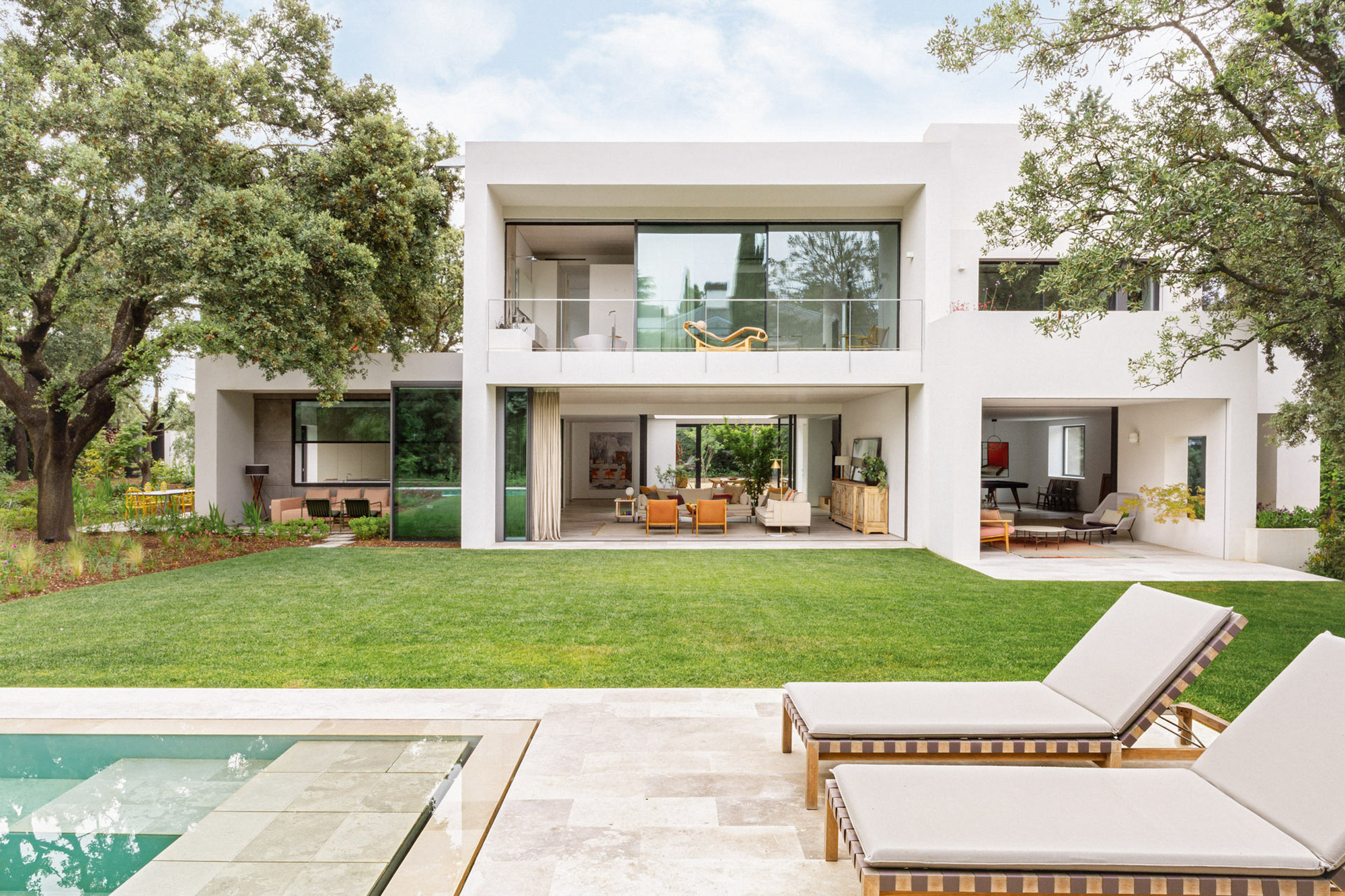White two-storey rear facade with open living rooms and glass balcony, seen across a lawn and travertine pool terrace with two lounge chairs
