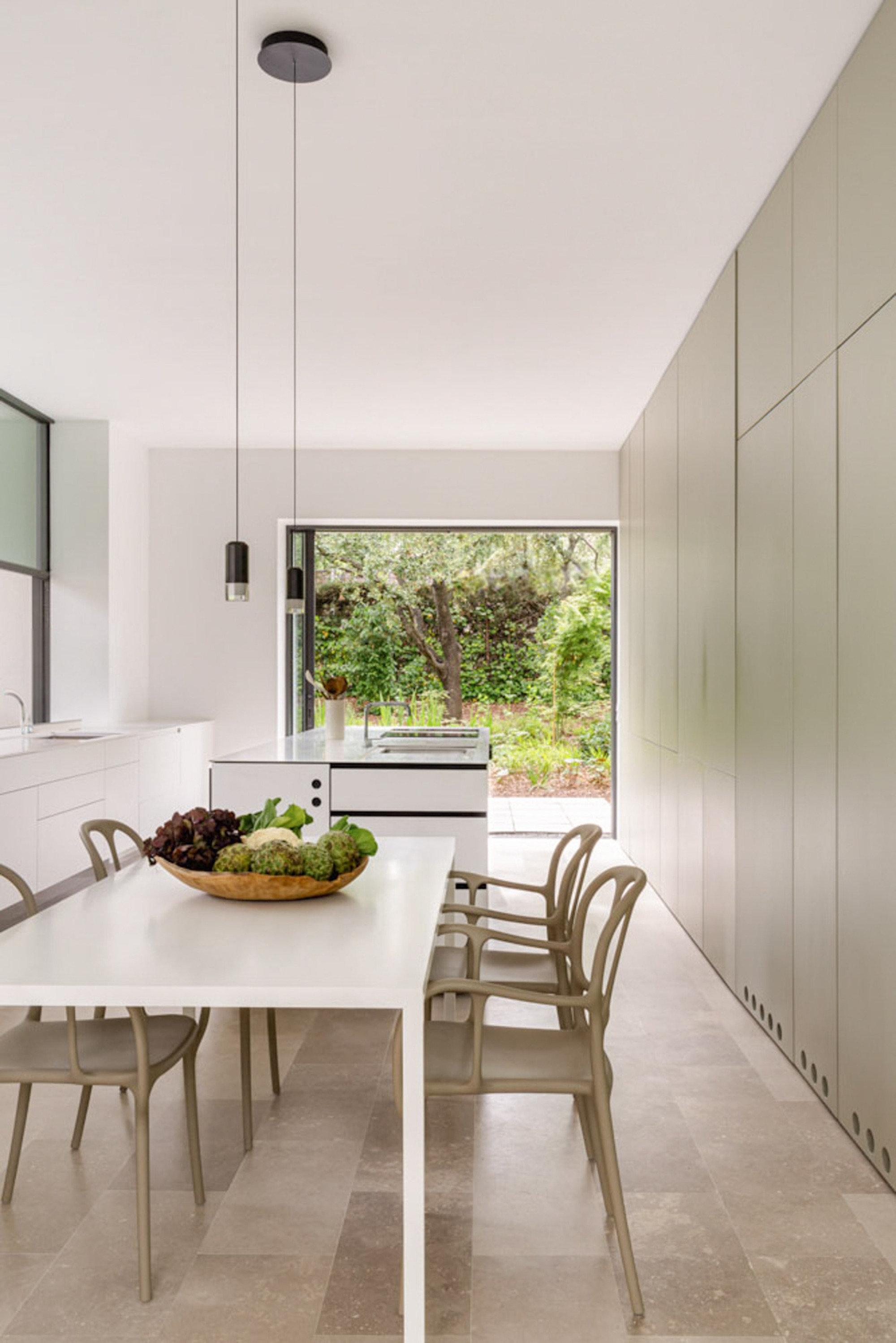 Kitchen and dining space with a white table, olive-toned chairs and tall cabinet wall, opening to a leafy garden