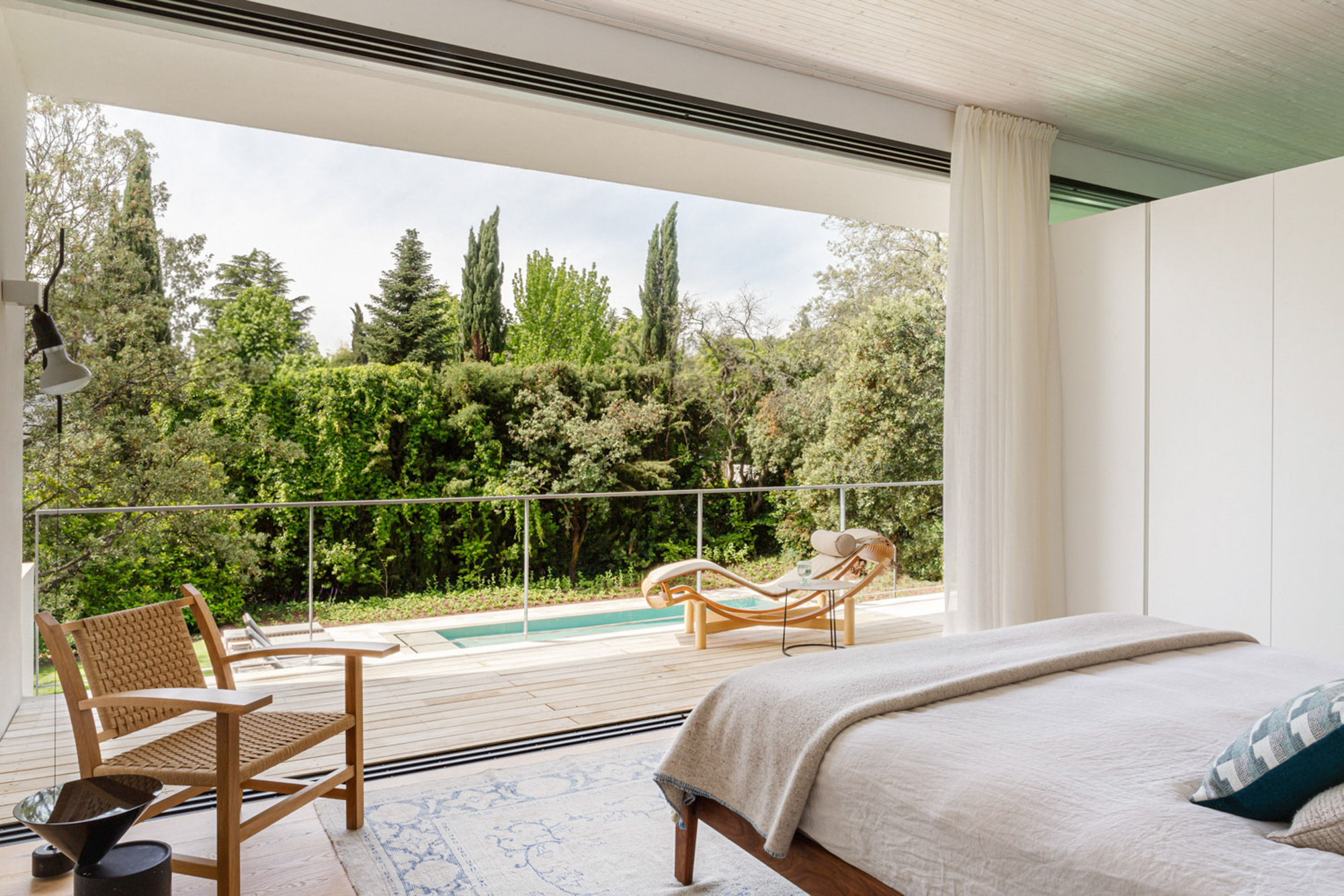 Bedroom with sliding glass wall opening to a timber deck, pool and a lounge chair, with a white curtain drawn aside