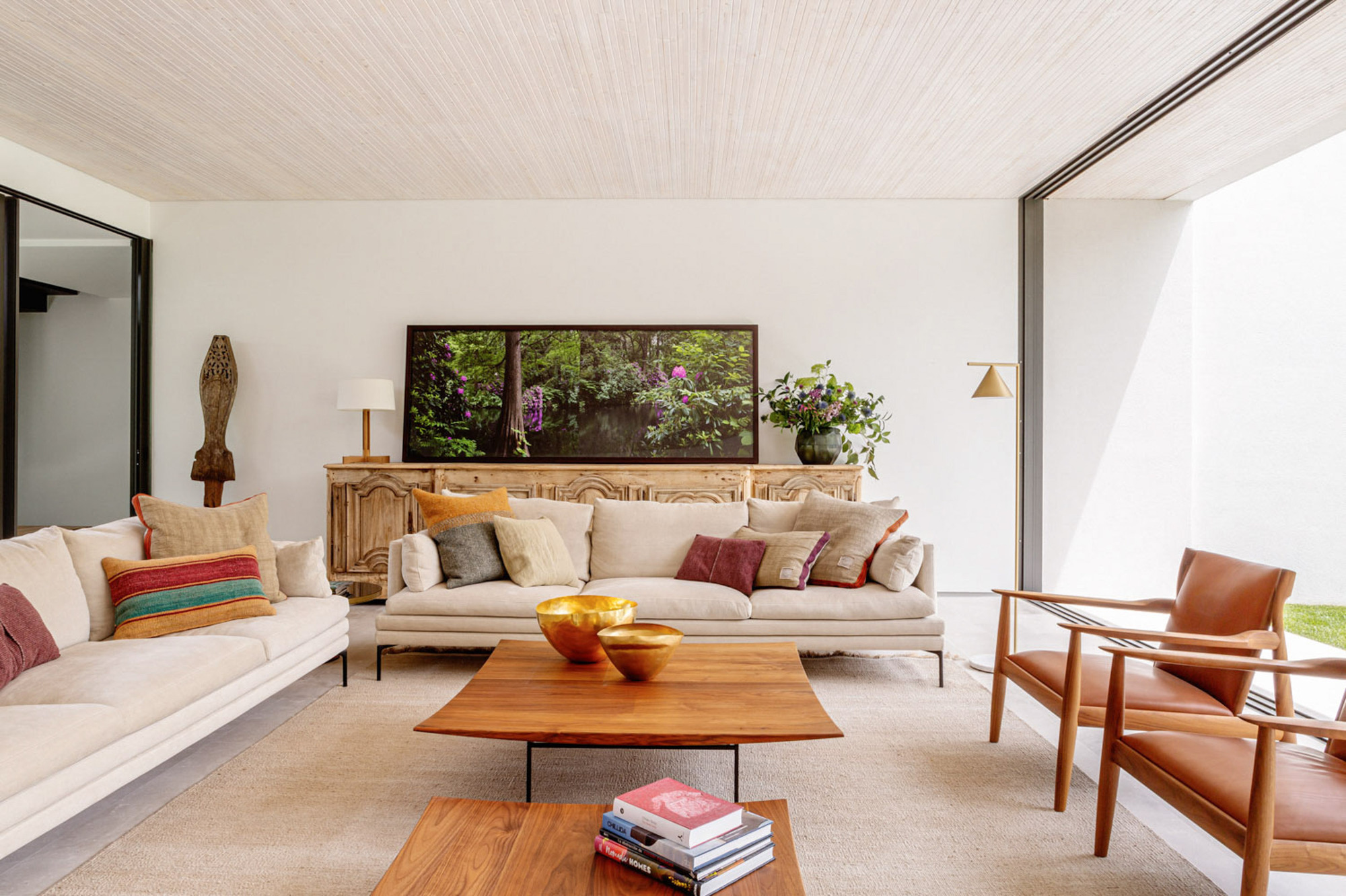 Living room with cream sofas, a walnut coffee table and leather armchairs facing a carved wooden sideboard, daylight entering from a side courtyard