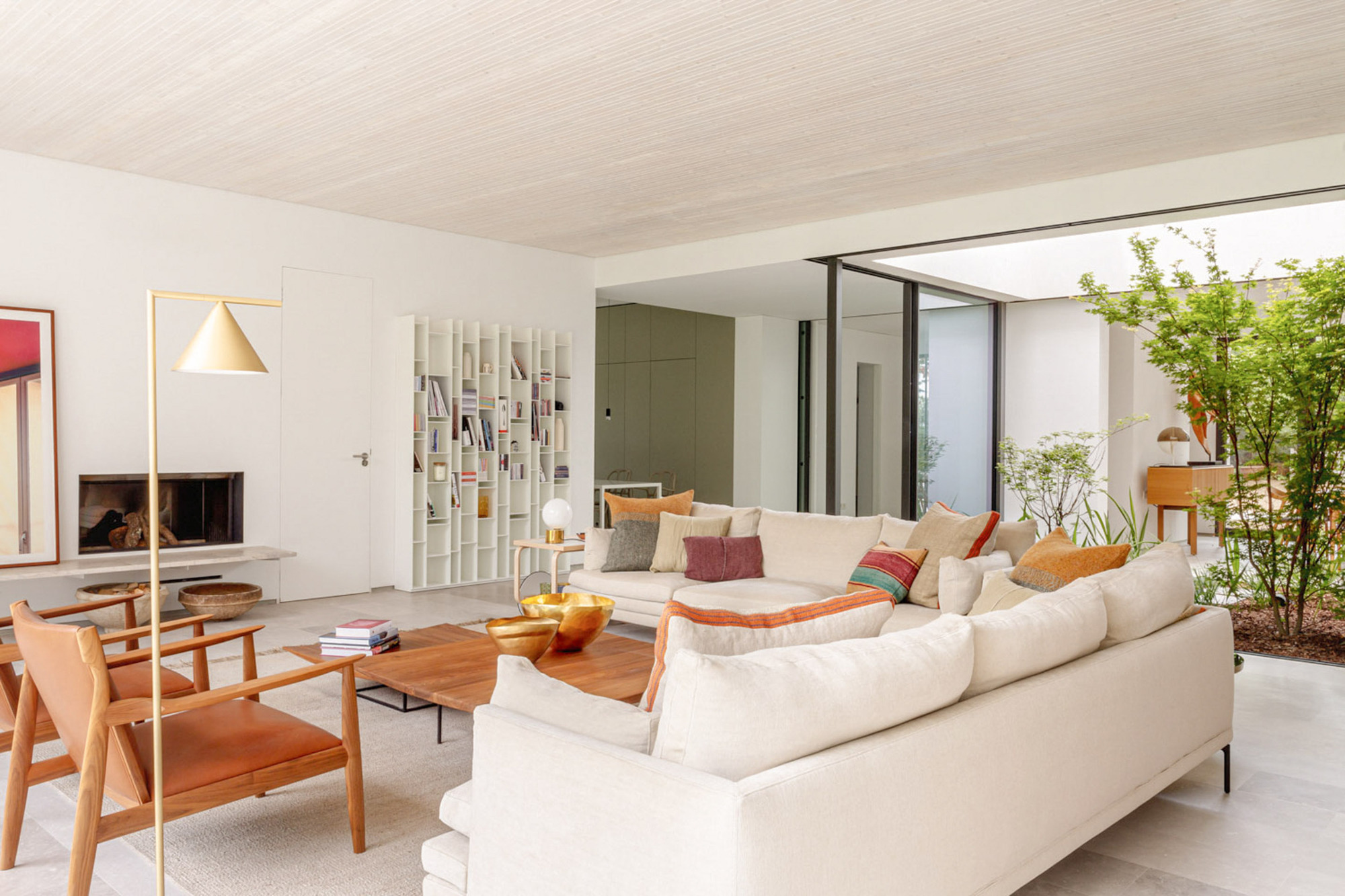 Corner sofa and tan leather chairs in an open living area with a white bookshelf and a small tree in the courtyard beyond