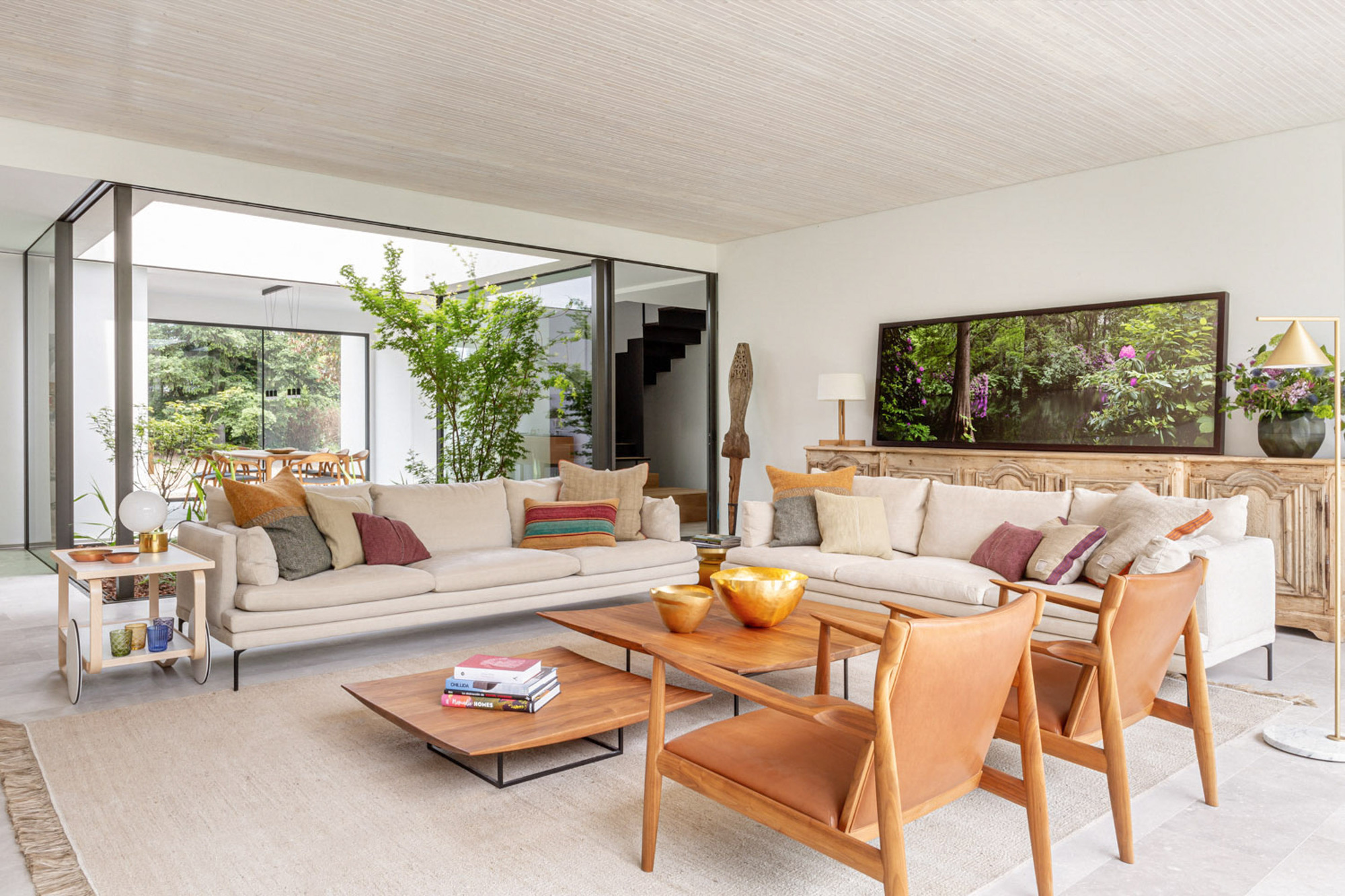 Two facing sofas and leather armchairs on a woven rug, with a sliding glass wall opening to a leafy courtyard