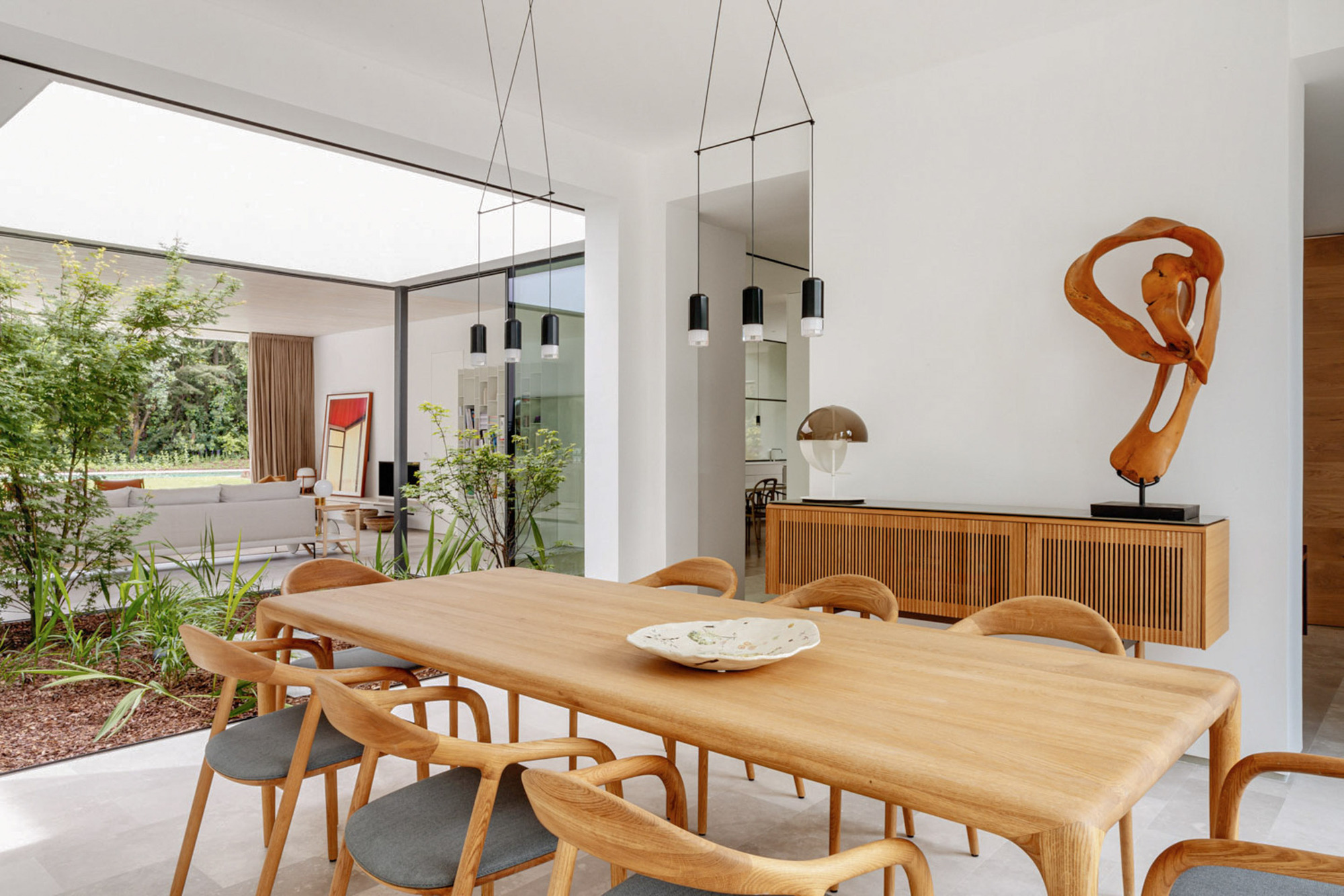 Oak dining table with curved chairs beside a slatted sideboard and wooden sculpture, a planted courtyard bed visible through the glass