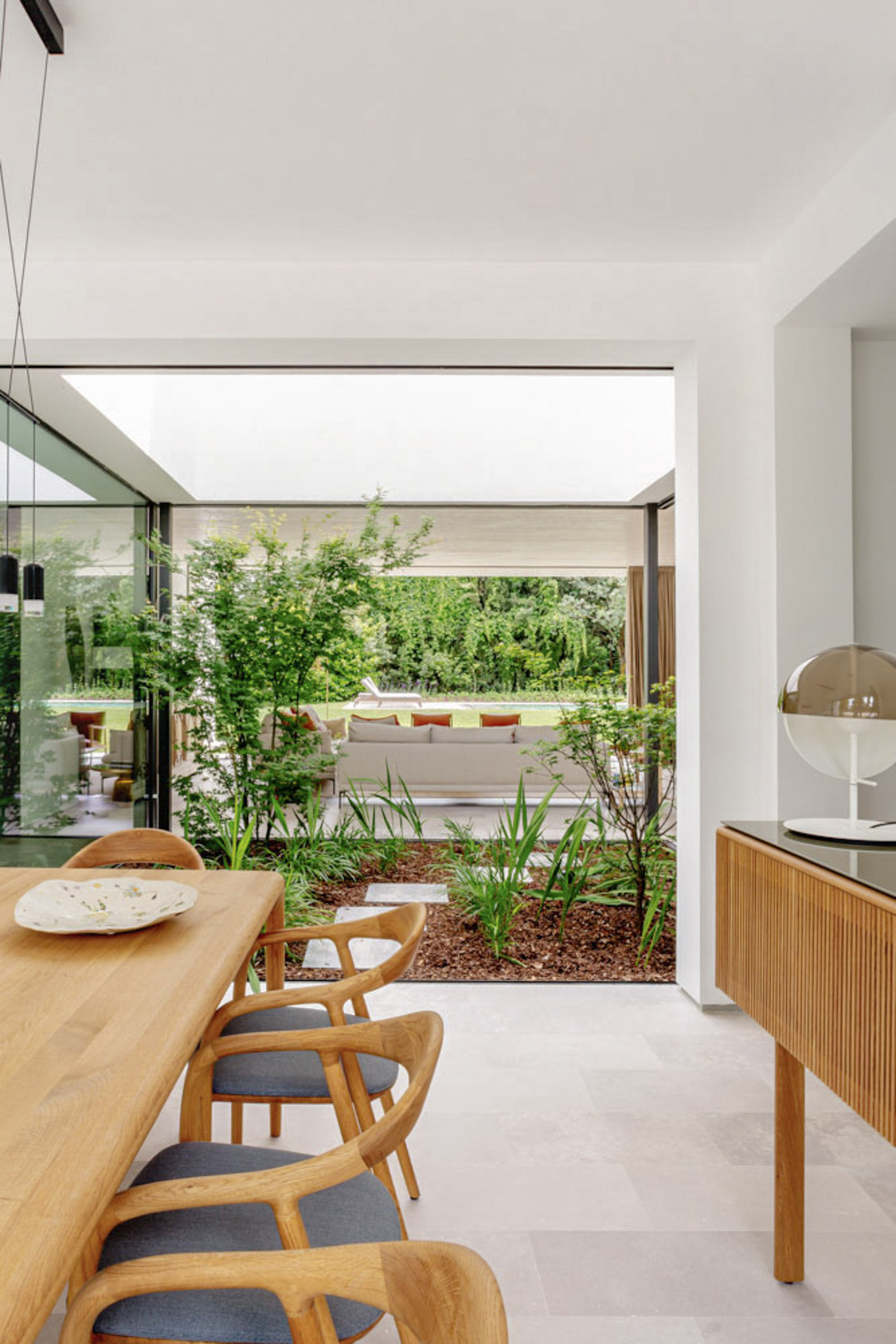 Dining table and oak chairs beside an open glass corner framing a planted courtyard bed with maples and grasses