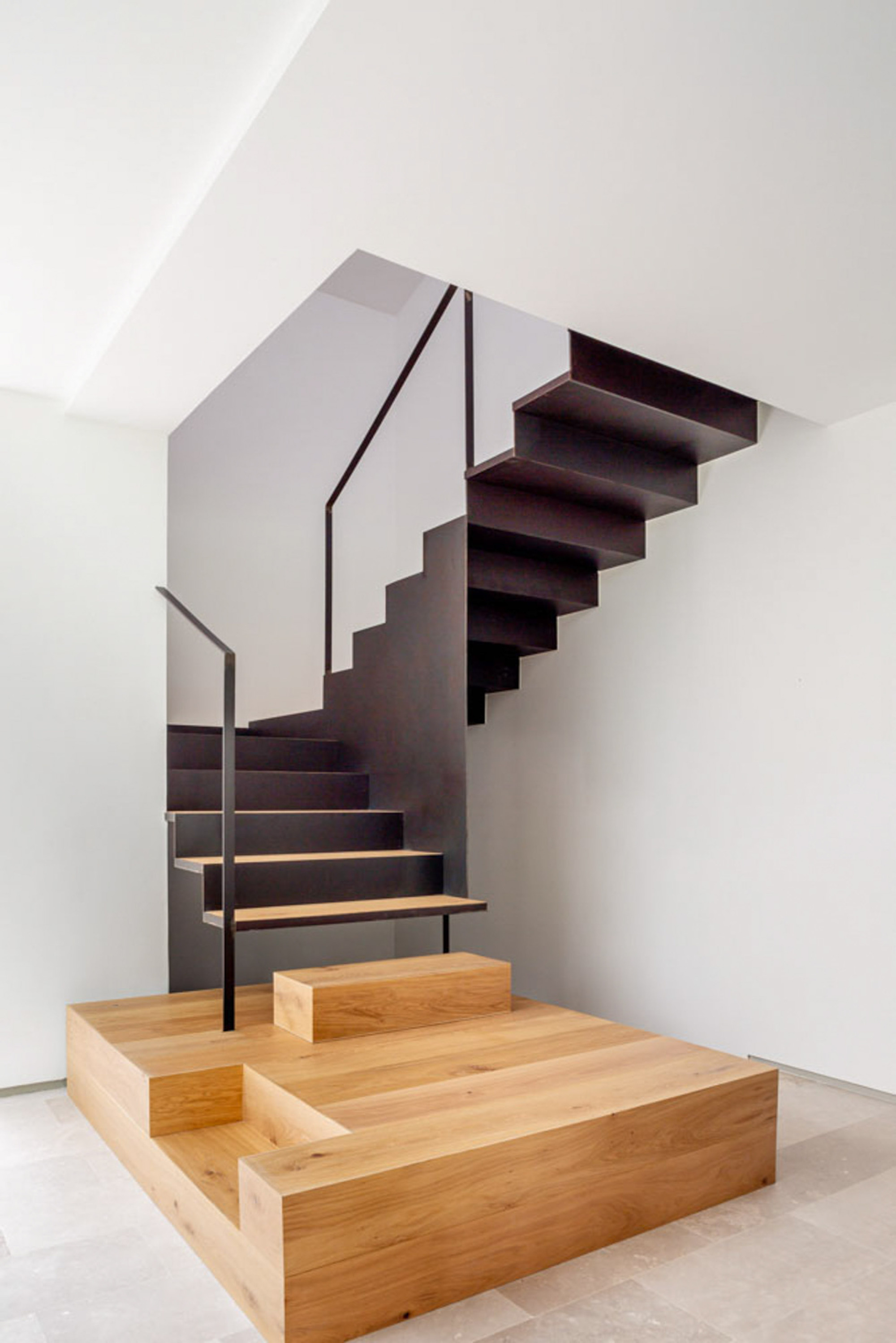 Black folded-steel staircase with oak treads rising from a stepped oak platform against white walls