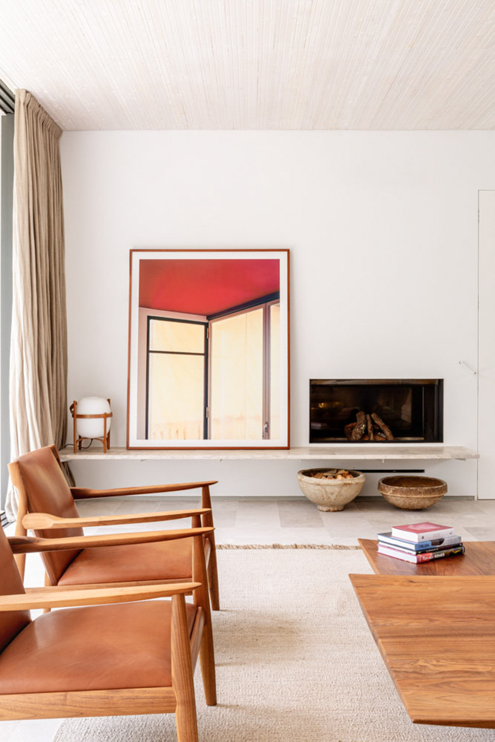 Low linear fireplace and stone ledge beneath a framed red-toned photograph, flanked by leather and timber armchairs in soft daylight