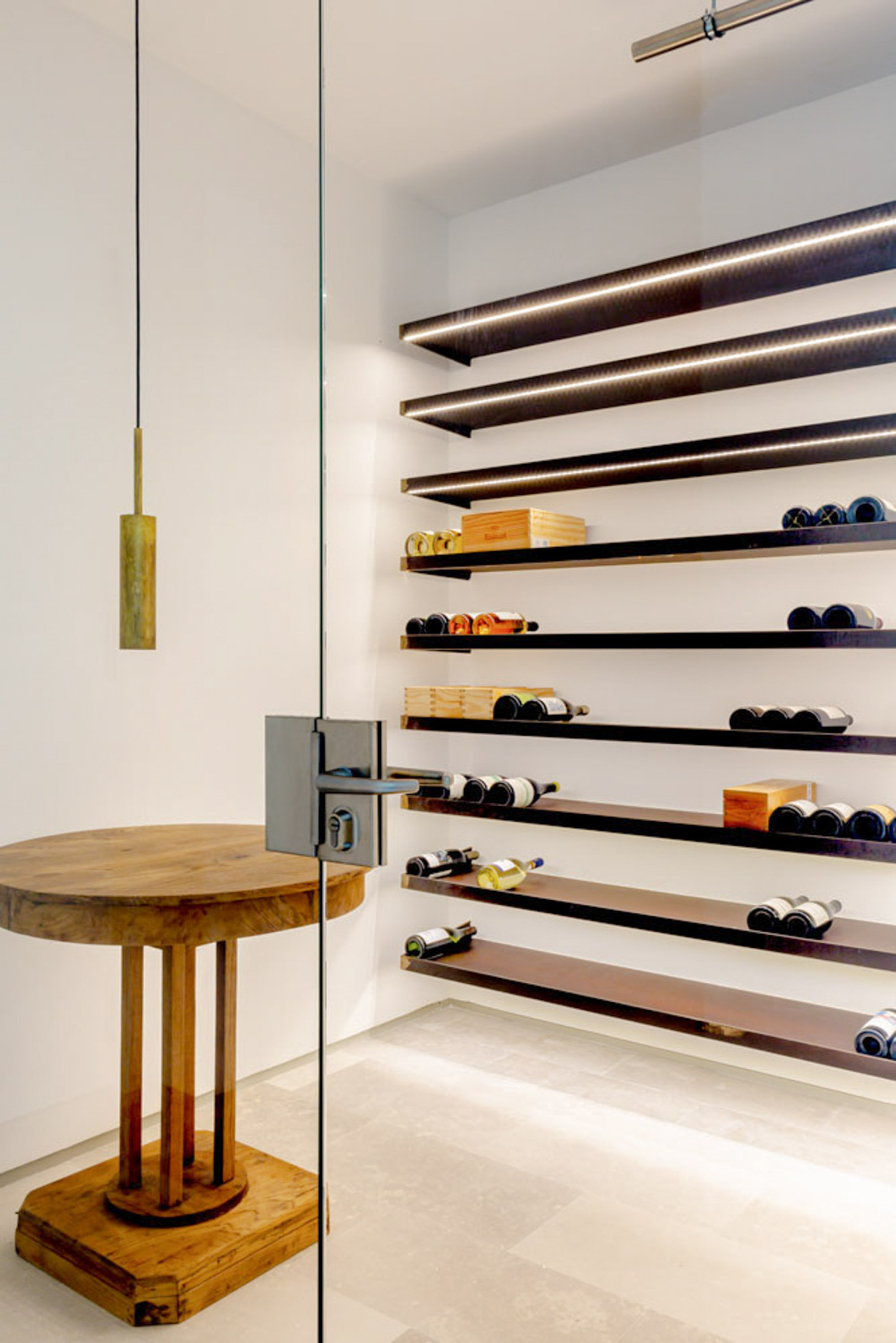 Glass-enclosed wine cellar with dark floating shelves lit by strip lights, beside a round timber pedestal table