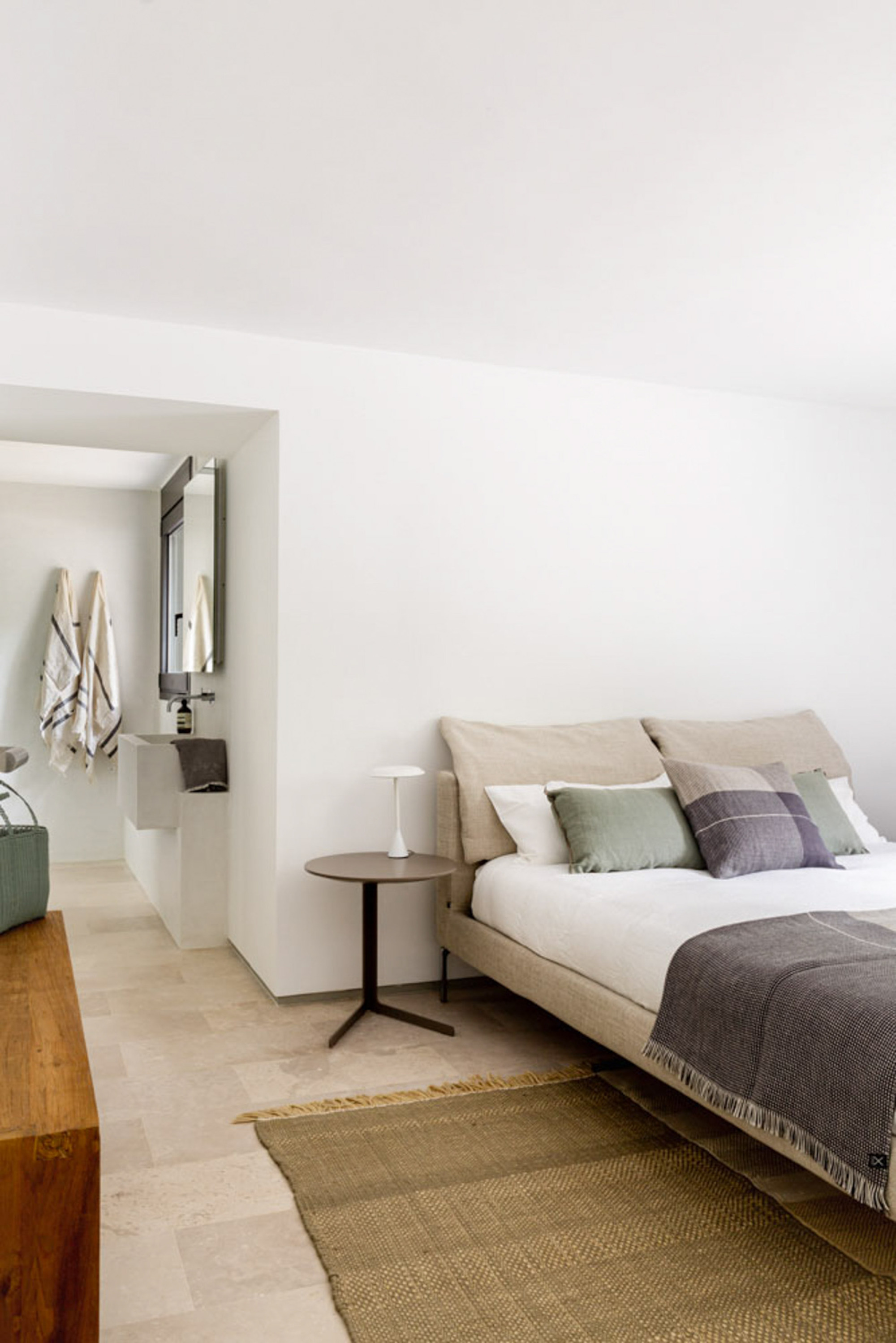 Bedroom with a low upholstered bed and woven jute rug, opening through a wall recess to a bathroom with hanging towels