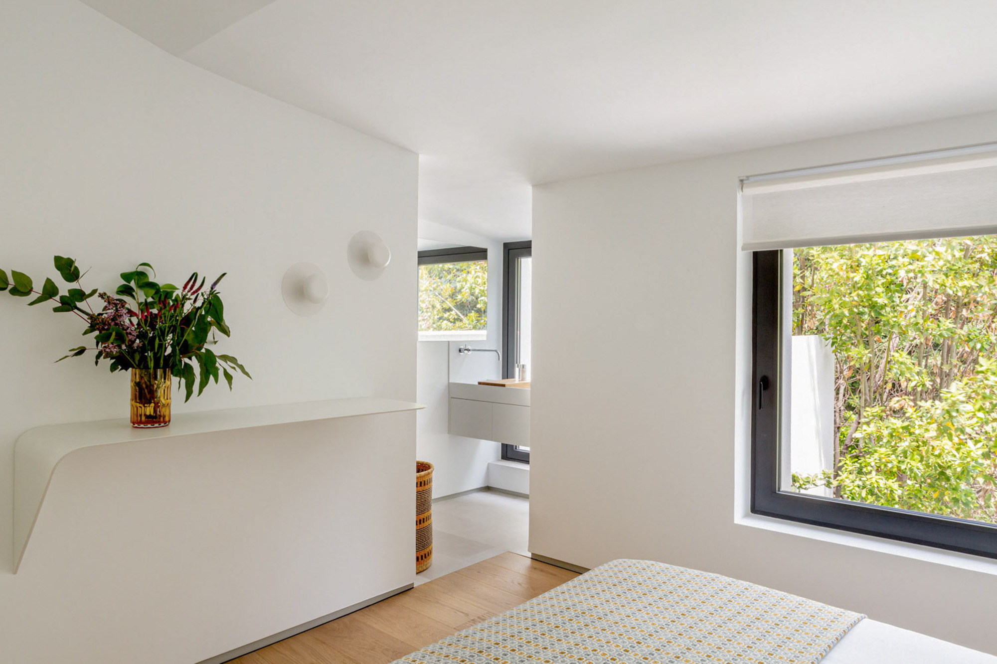 Bedroom corner with a curved white wall shelf holding a flower vase, and a window framing green foliage