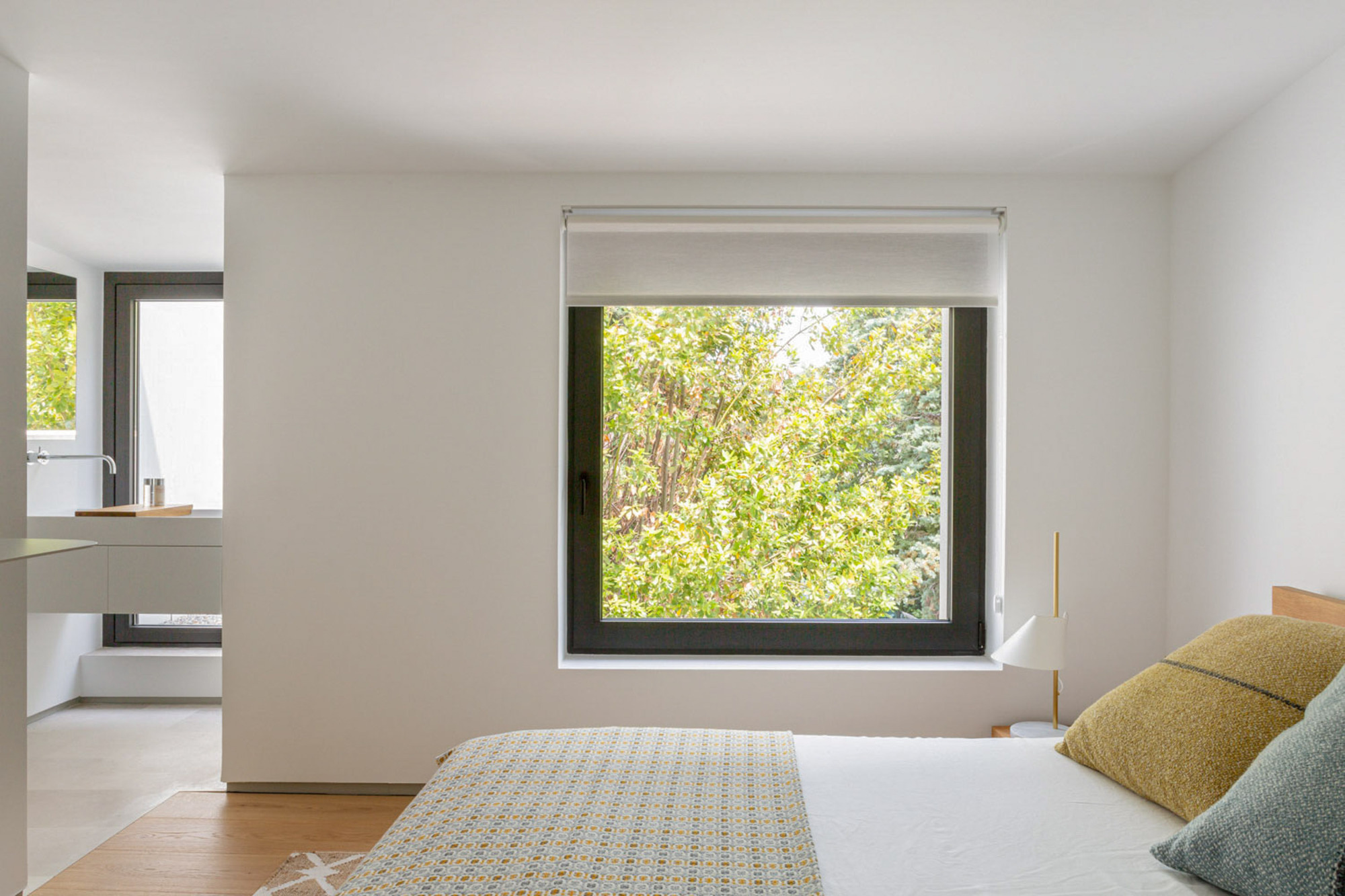 Bed facing a square black-framed window with a roller blind, leafy tree canopy outside and an open bathroom to the left