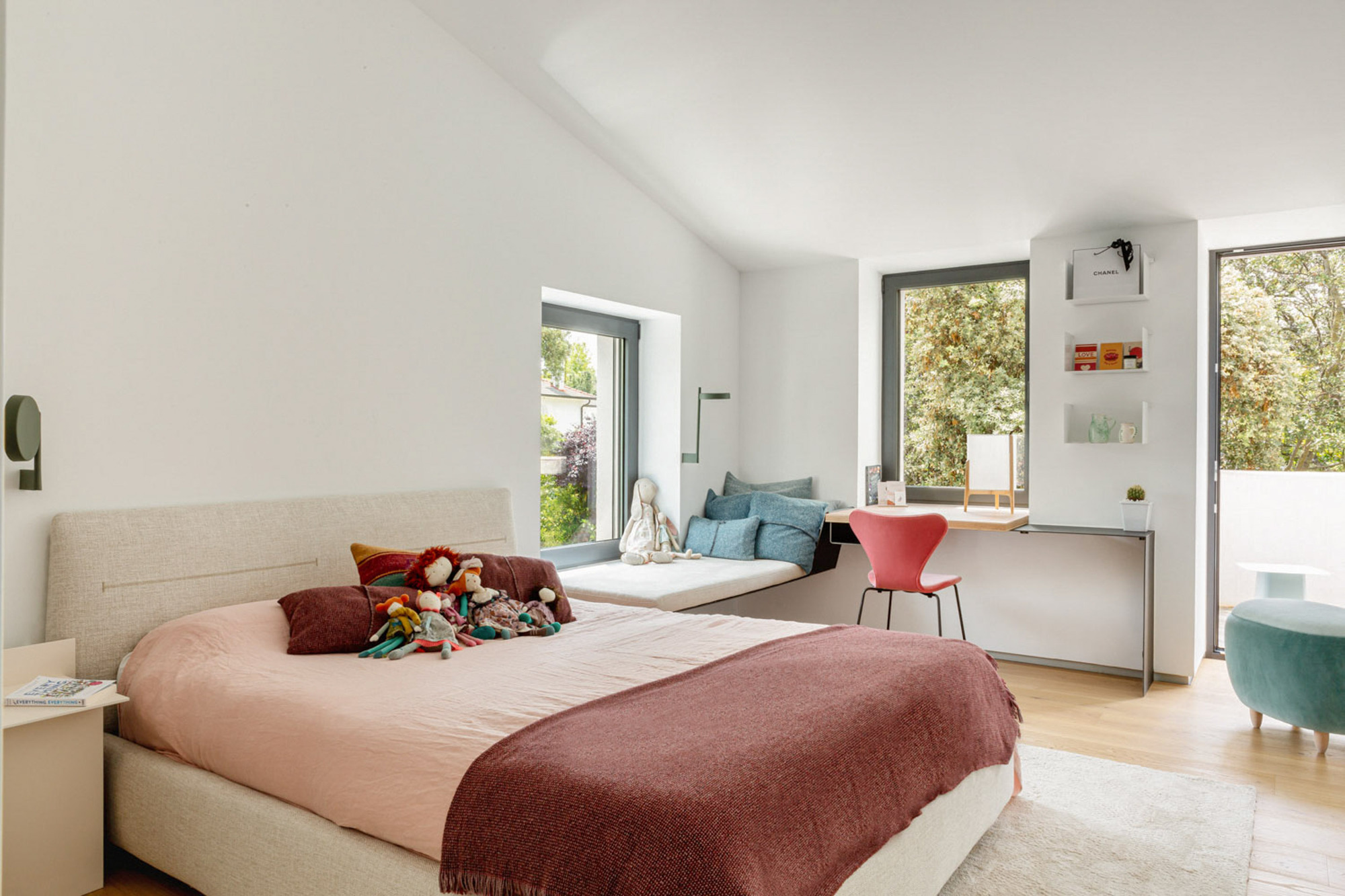 Child's bedroom under a sloped ceiling with a window seat, built-in desk, red chair and balcony door opening to treetops