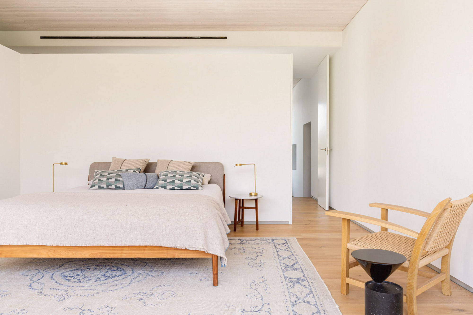 Bedroom with a low timber bed, pale blue rug and woven lounge chair beside a white partition wall in soft daylight