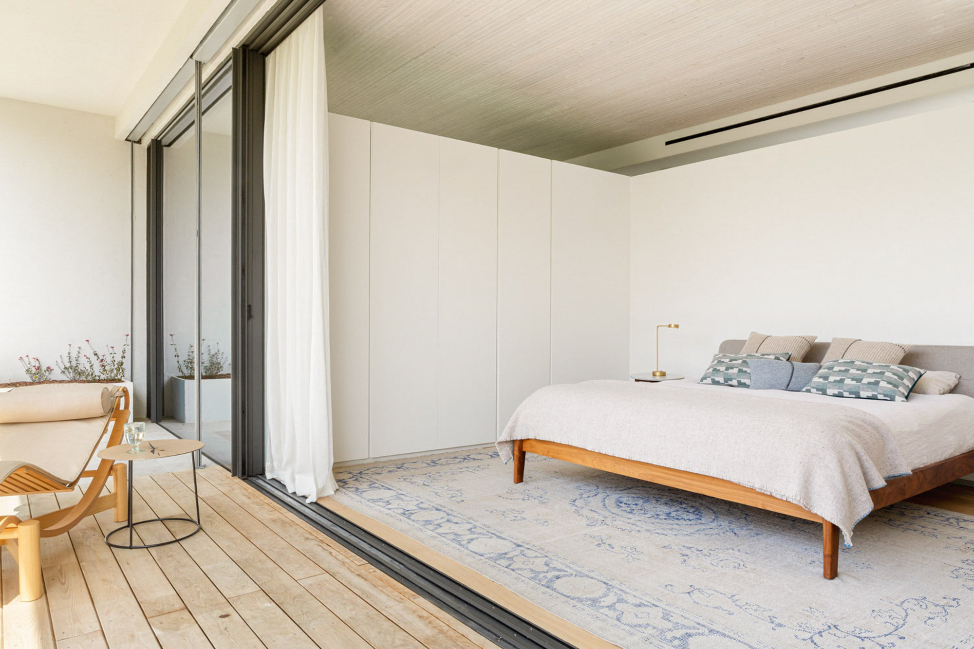 Bedroom opening through a sliding glass wall and white curtain onto a timber deck with a lounger in afternoon light