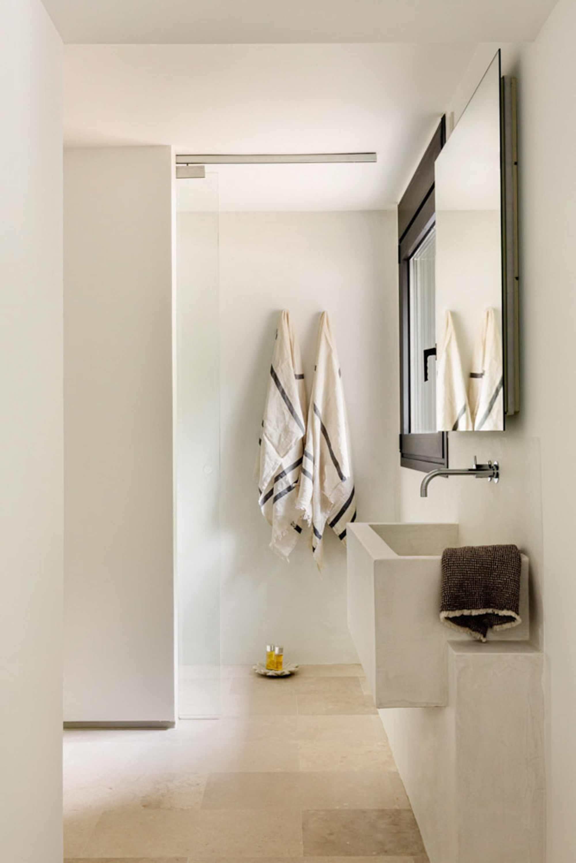 Bathroom with a concrete wall-hung basin, striped linen towels and a glass shower screen over pale limestone floor tiles