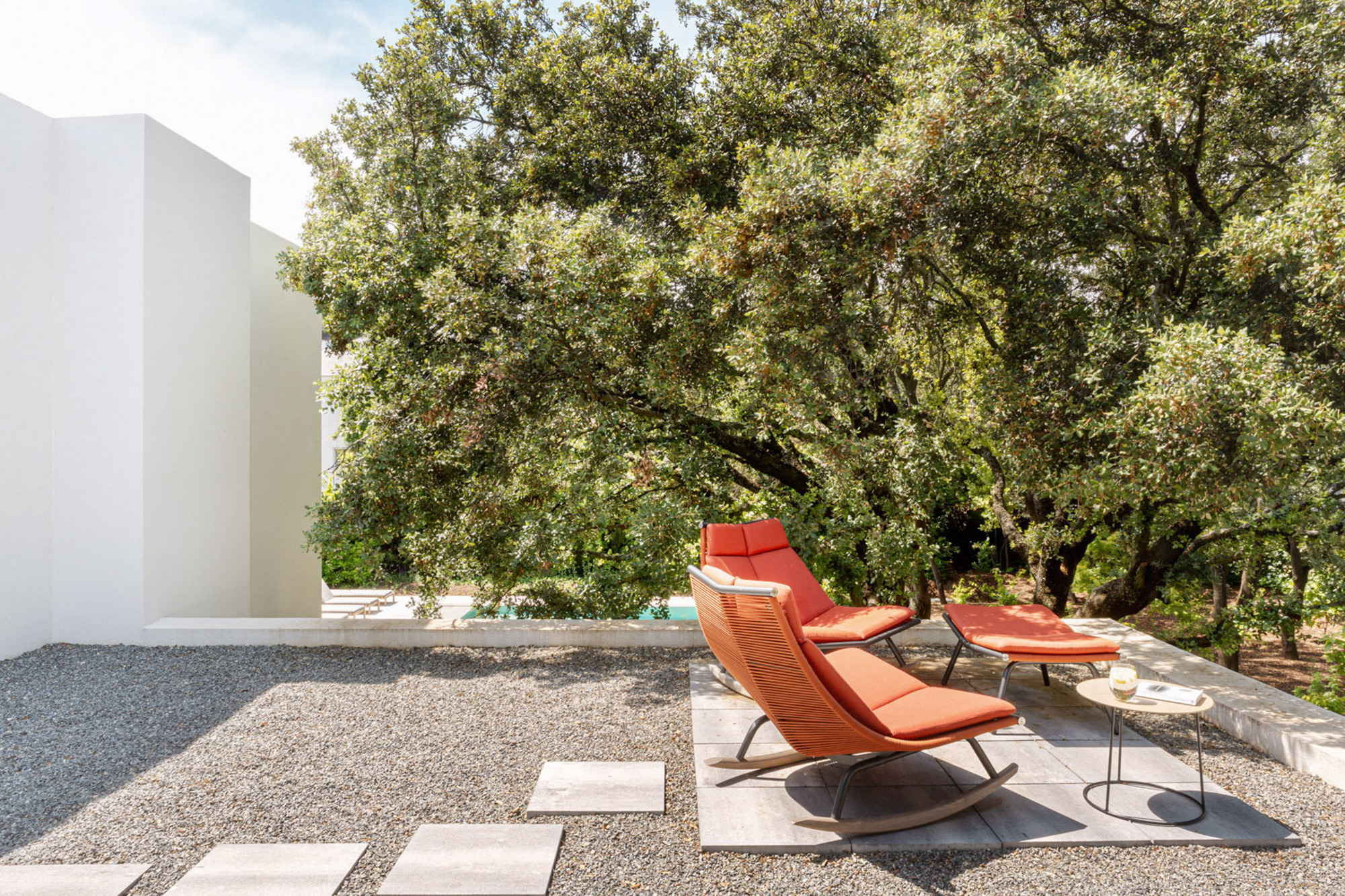 Two orange rocking loungers on a paved pad in a gravel terrace beneath a large oak tree in bright sun