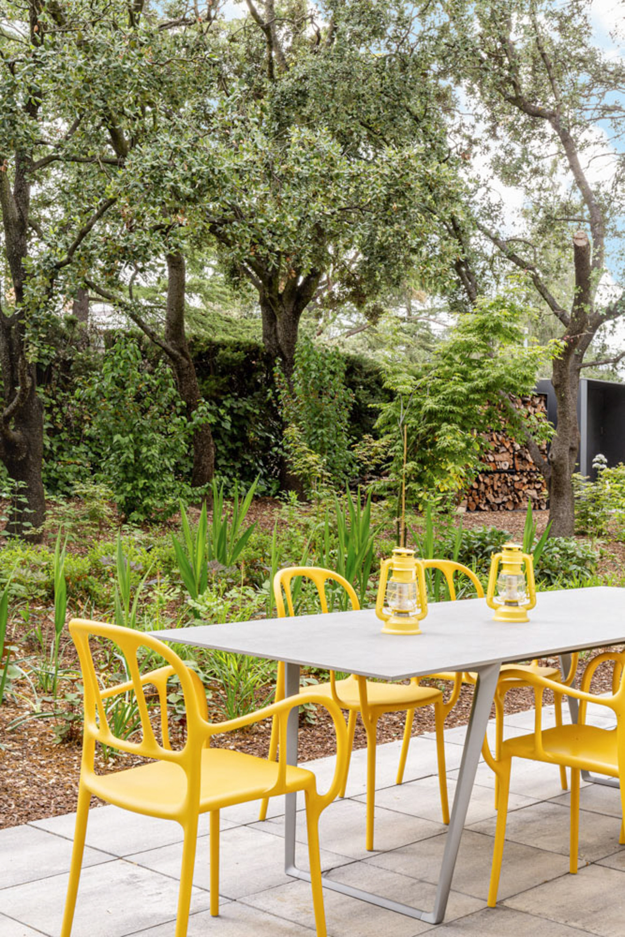 Close view of a grey-topped table with yellow chairs and two lanterns, with a stacked log pile behind in dappled shade