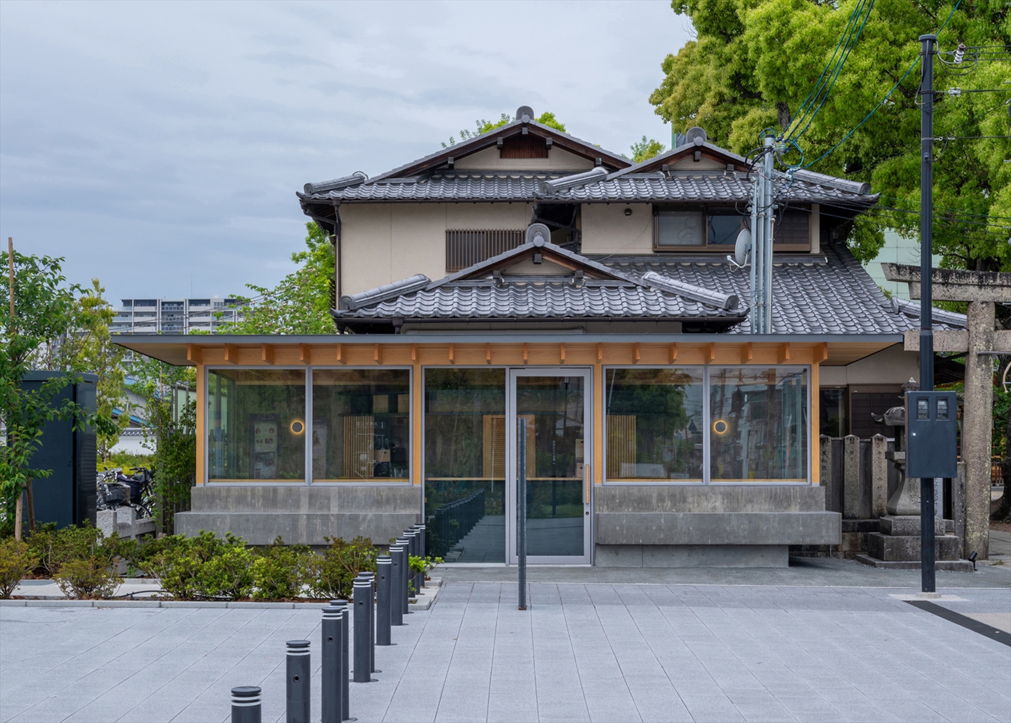 Front view of a small timber-eaved kiosk with concrete base and glazed walls in front of a tiled-roof house, under overcast sky