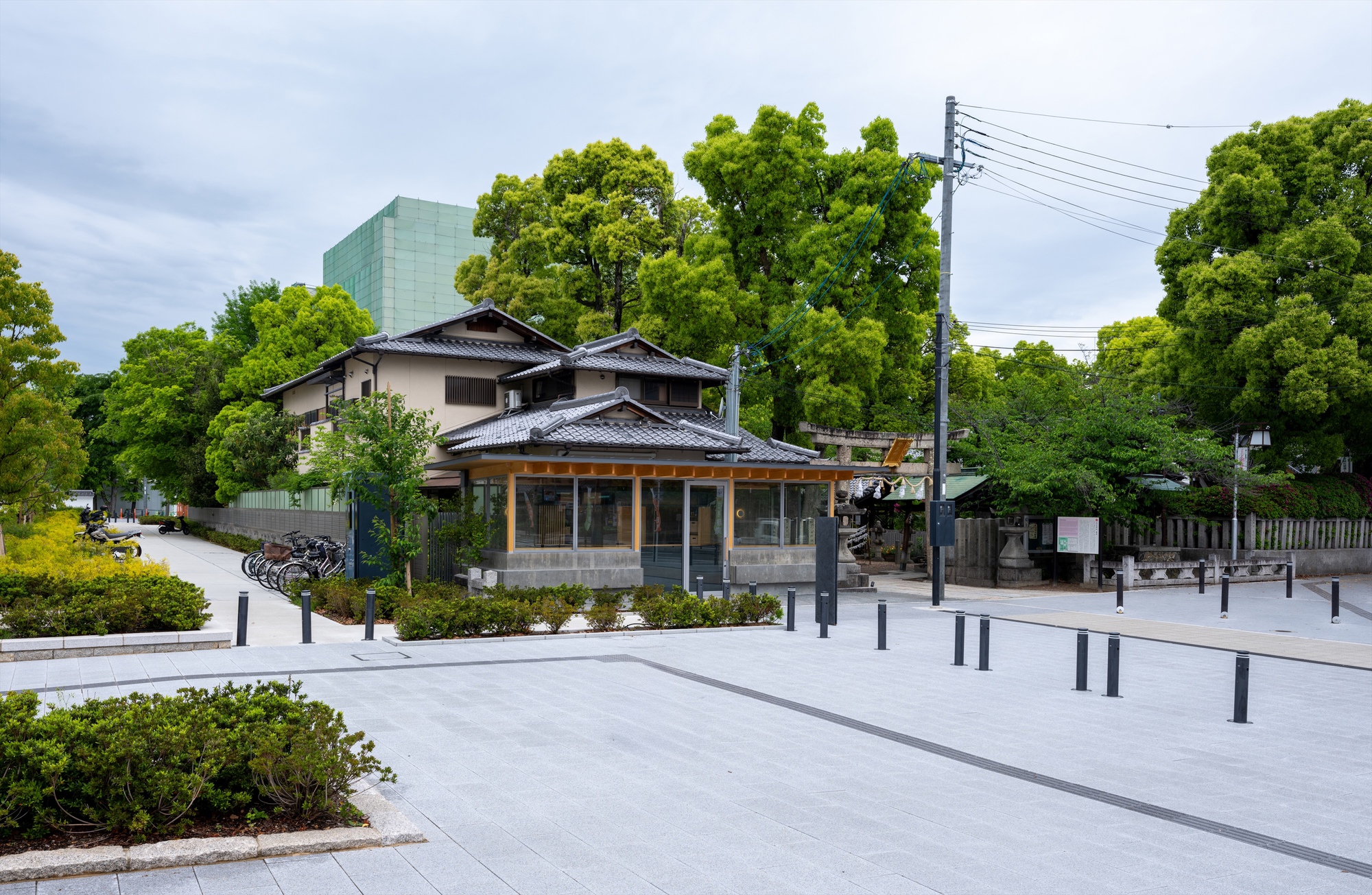 Wide view of the glazed kiosk beside a tiled-roof house, with bollards across the paved plaza and tall green trees